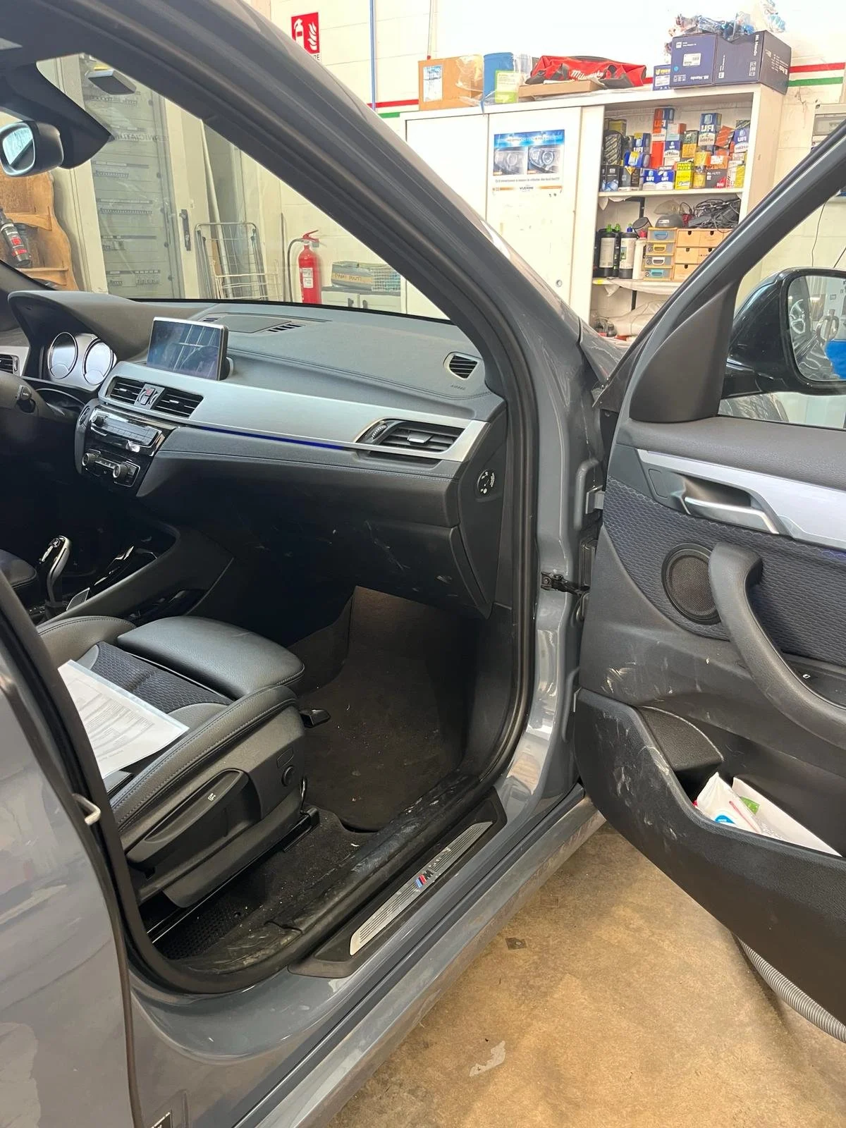 Interno di un'auto sportiva di colore grigio, vista dal lato aperto della portiera del conducente, con pannelli e sedili di colore grigio, in un garage con scaffali di attrezzi e scatole sullo sfondo.