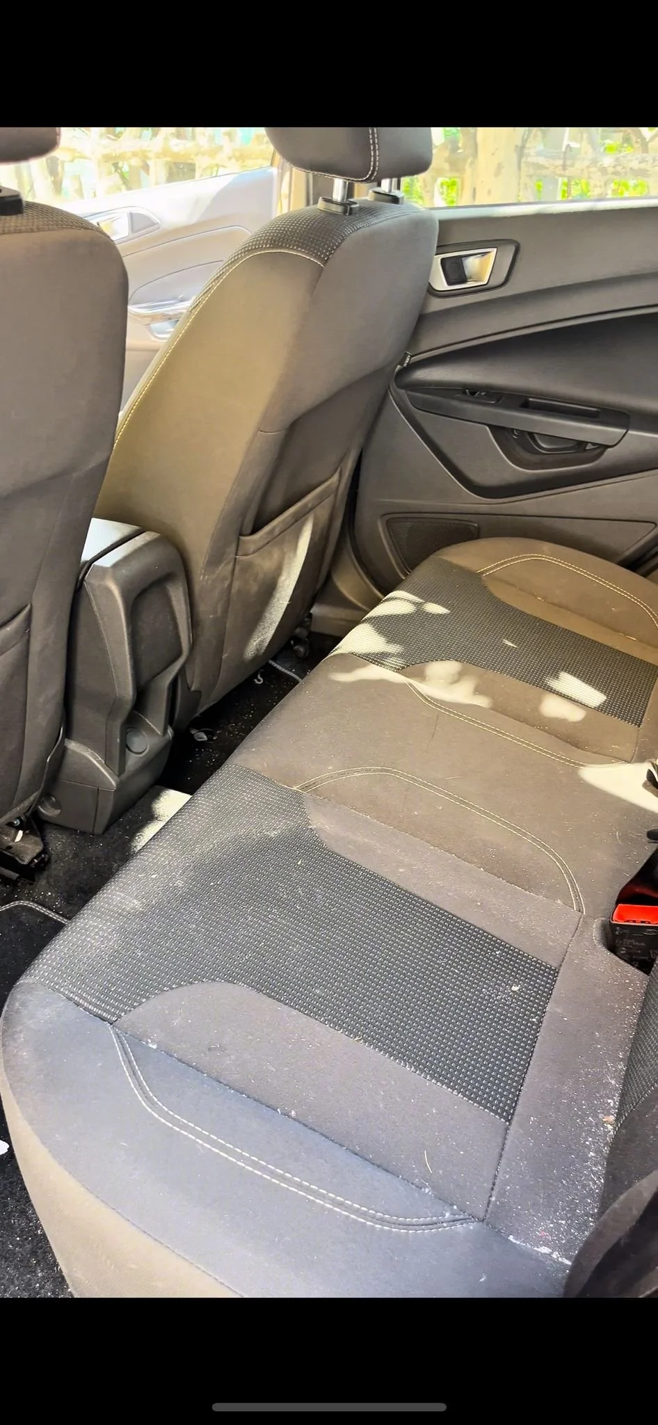 L'interno di un'auto, vista dal sedile posteriore, con sedili in tessuto nero, alcuni segni di sporco e polvere, e portiere con maniglie nere e dettagli in plastica.