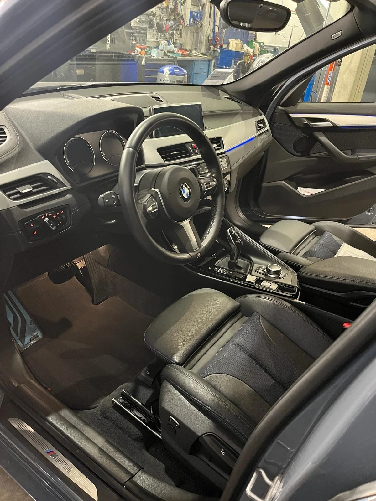 Interno di un'auto BMW con volante, sedili in pelle e plancia moderna, di fronte a un garage con attrezzi.