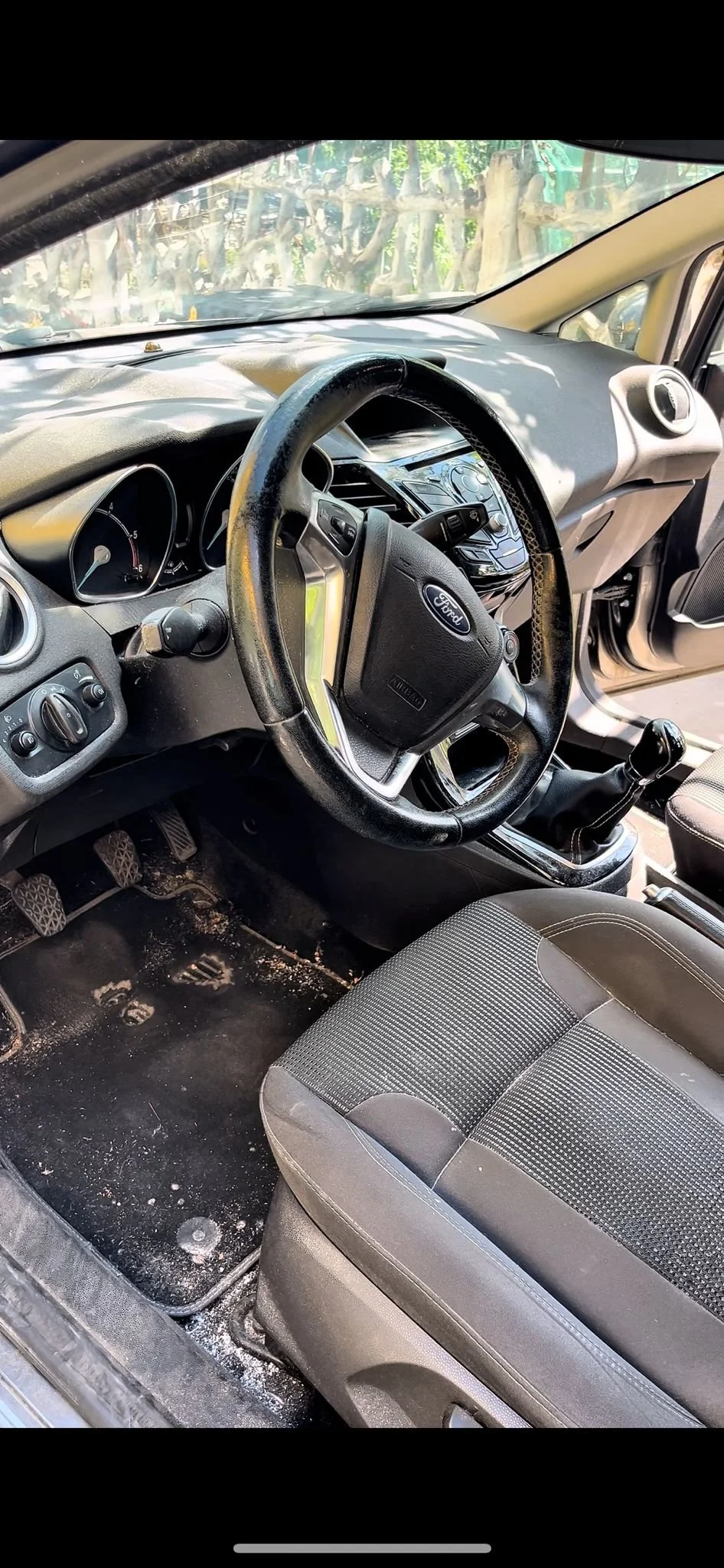 Interno di un'auto Ford con il volante e il cruscotto visibili, sedile con tessuto a scacchi, tappetini sporchi e comandi della vettura.