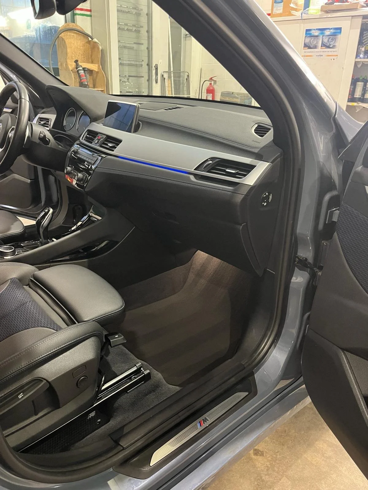 Interno di un'auto sportiva con volante, cruscotto, display, e sedili in pelle. Logo M sulla soglia.