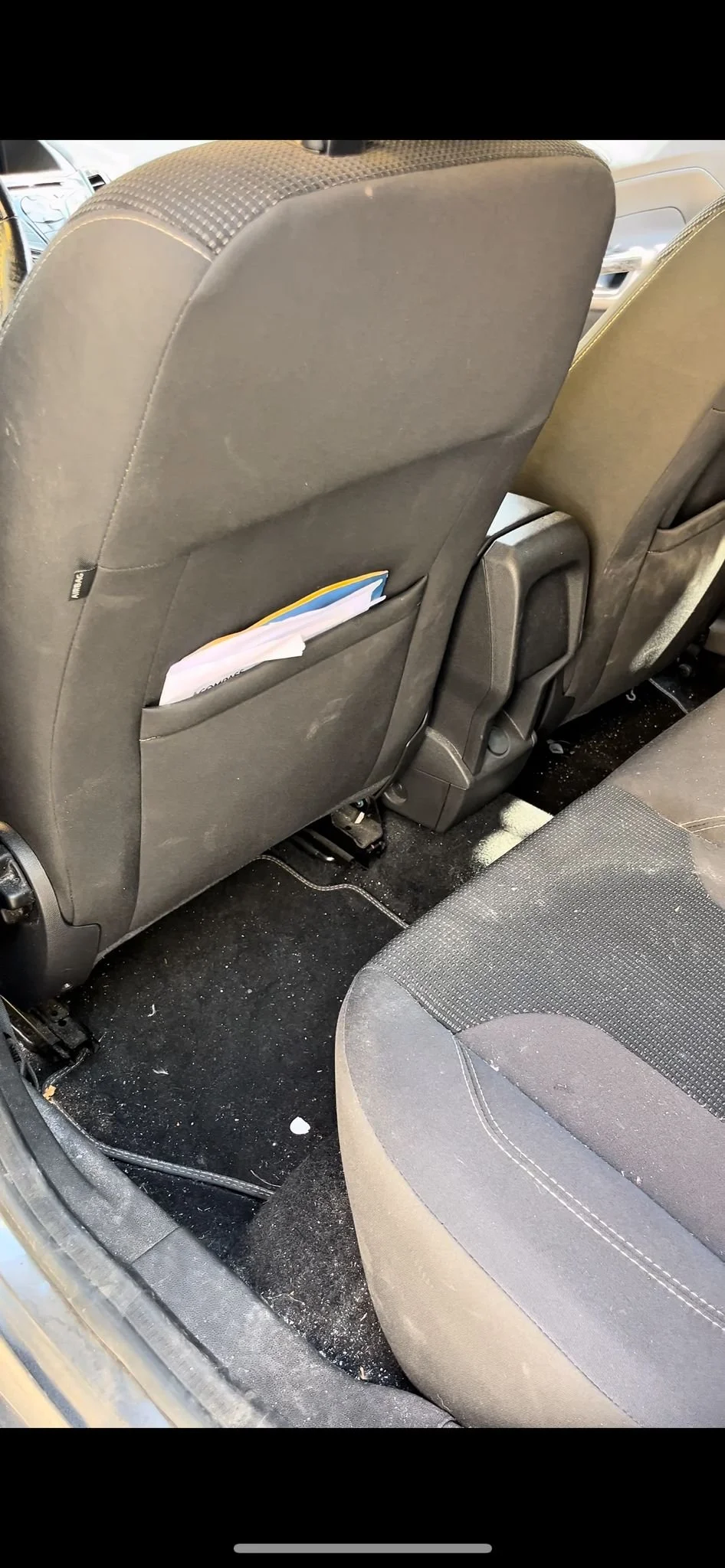 Interno di un'auto con sedili neri, pavimento sporco e sacca portaoggetti con documenti nella parte posteriore del sedile anteriore.