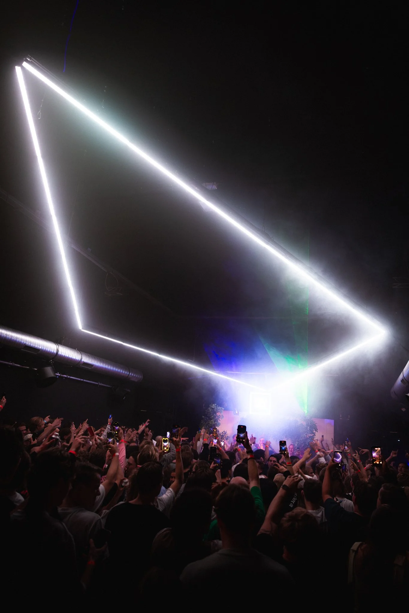 A large crowd of people attending a concert or a DJ event, facing a bright stage with laser lights forming geometric patterns overhead.