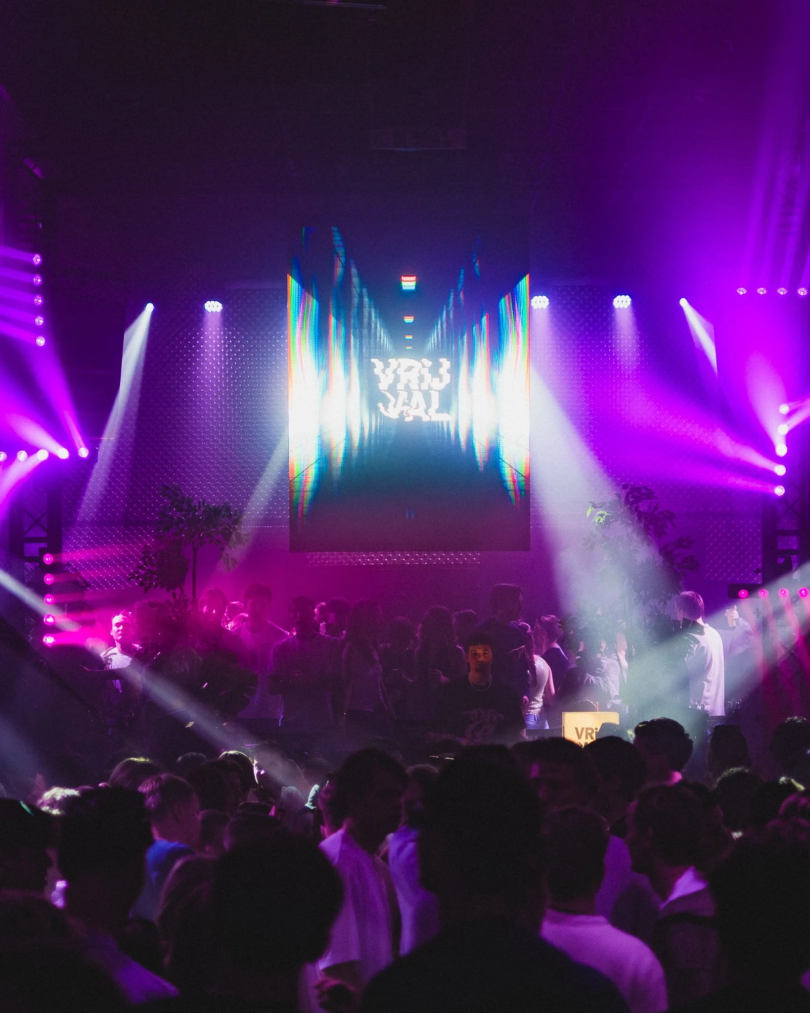 Crowd of people at an electronic music concert with colorful stage lights and a large screen displaying distorted visuals.