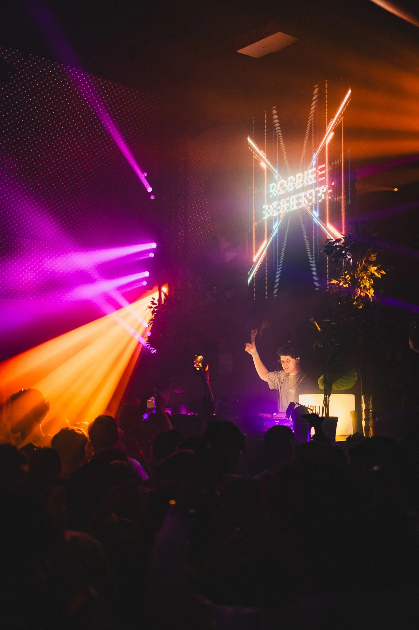 A DJ performs at a nightclub with colorful neon lights and laser beams, a crowd enjoying the music, and a digital sign reading 'Do Not Stop' in the background.