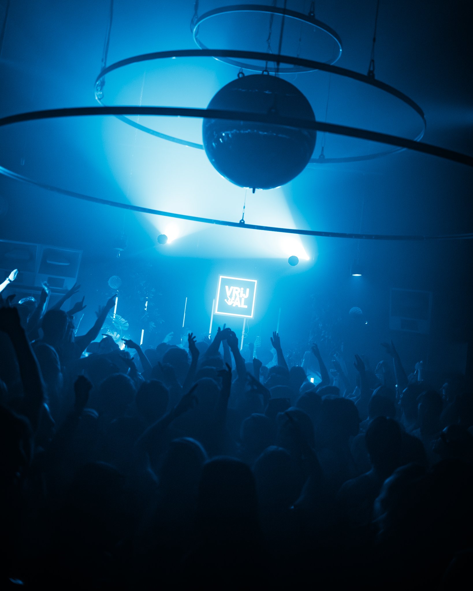 Crowd dancing at a nightclub or concert with blue lighting and a sign that says 'VRIJ SAL'.