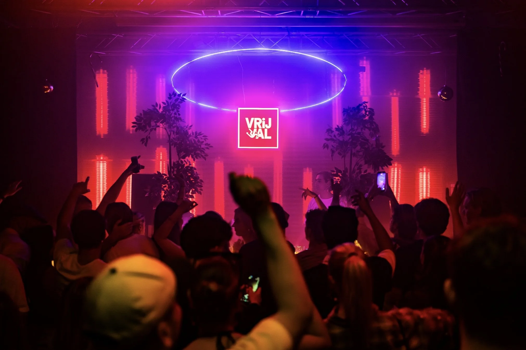 A crowded indoor concert with a stage, vibrant purple and red neon lights, and a sign that reads 'VRIJ VAL,' with audience members raising their hands and taking photos.