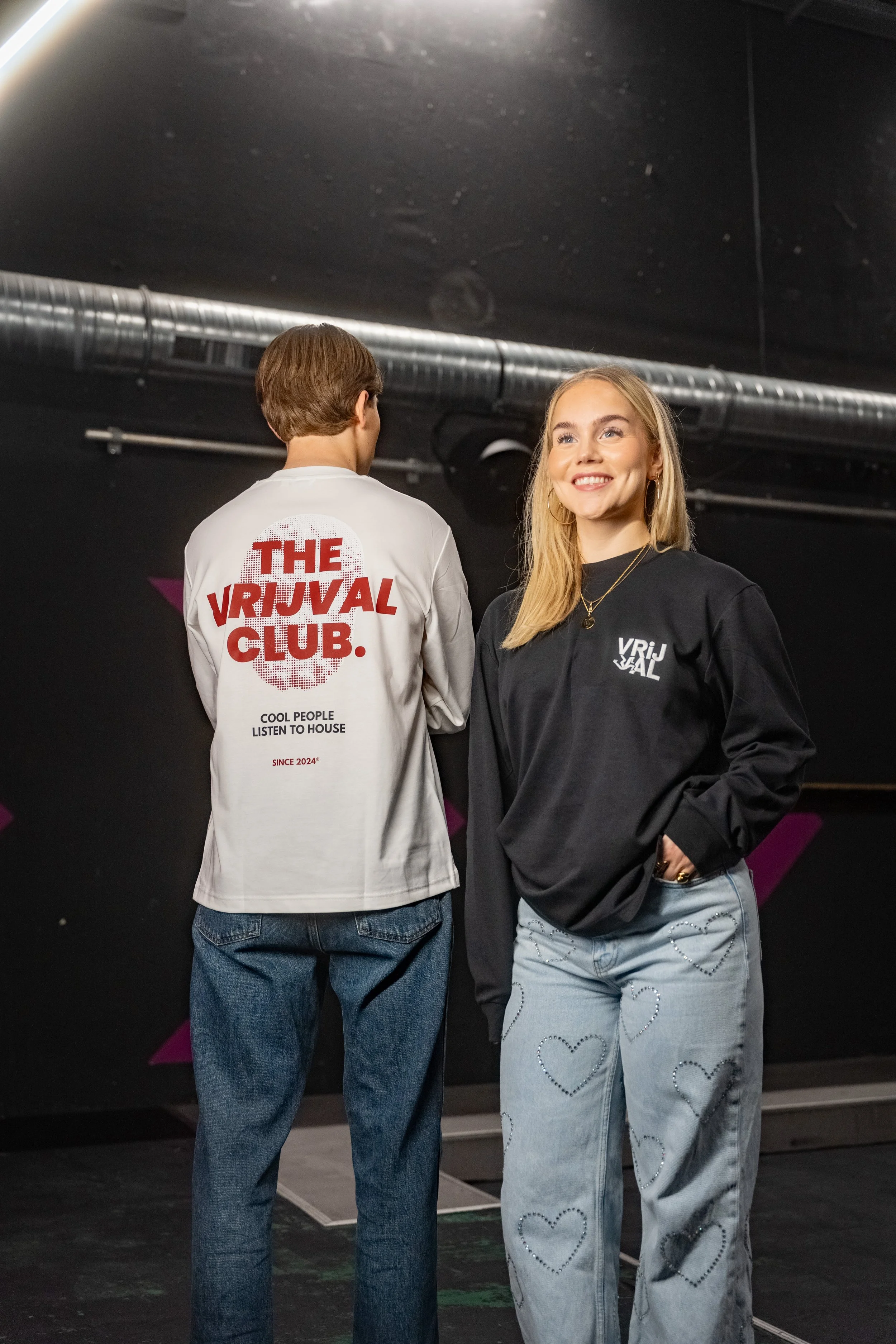 Two young people standing in a dark room, facing away from each other, wearing casual clothing. The person on the left has a white shirt with red text on the back that reads "The VIRIUAL CLUB" and the person on the right is wearing a black sweatshirt