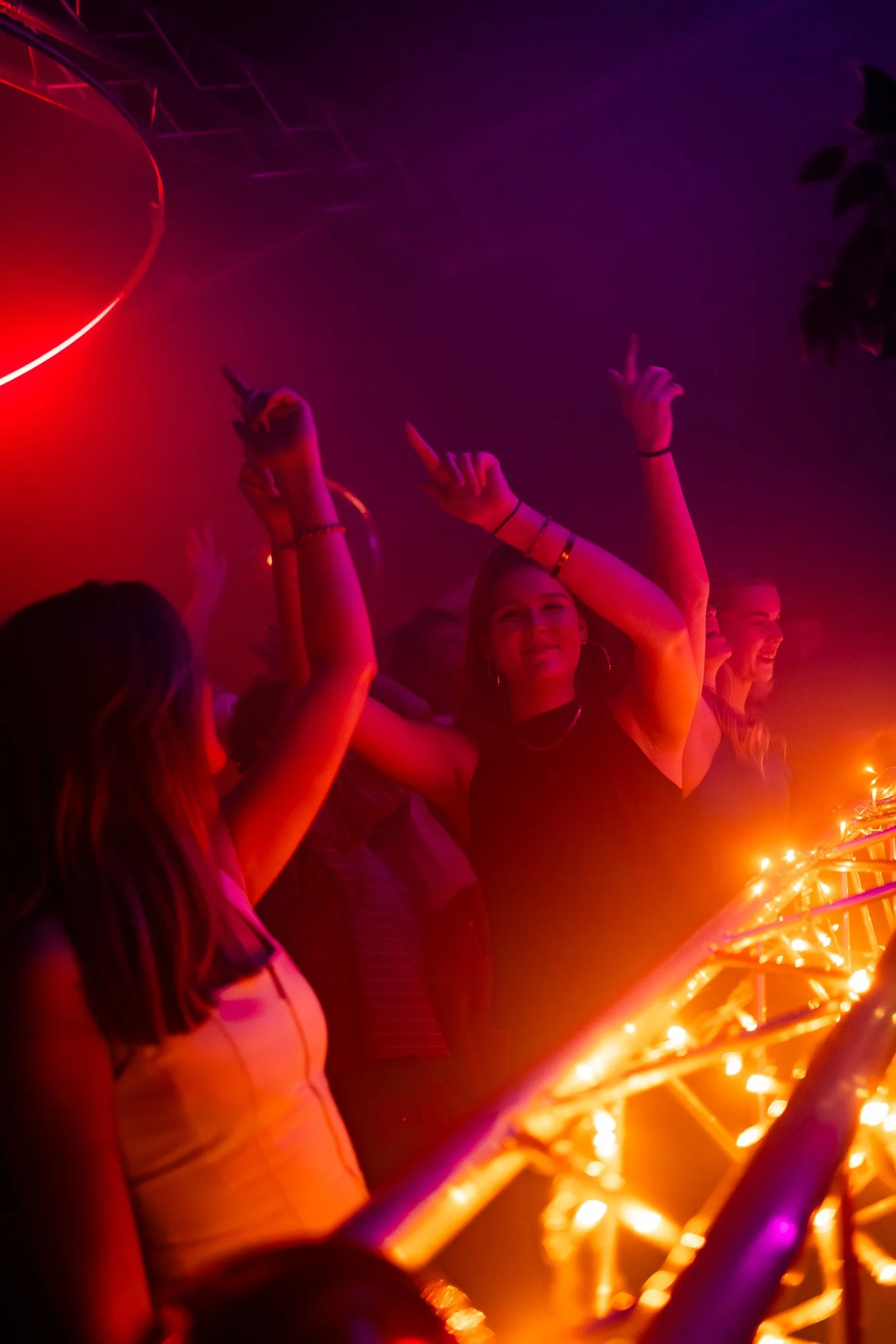 People dancing at a party or nightclub with colorful lights and a DJ booth.