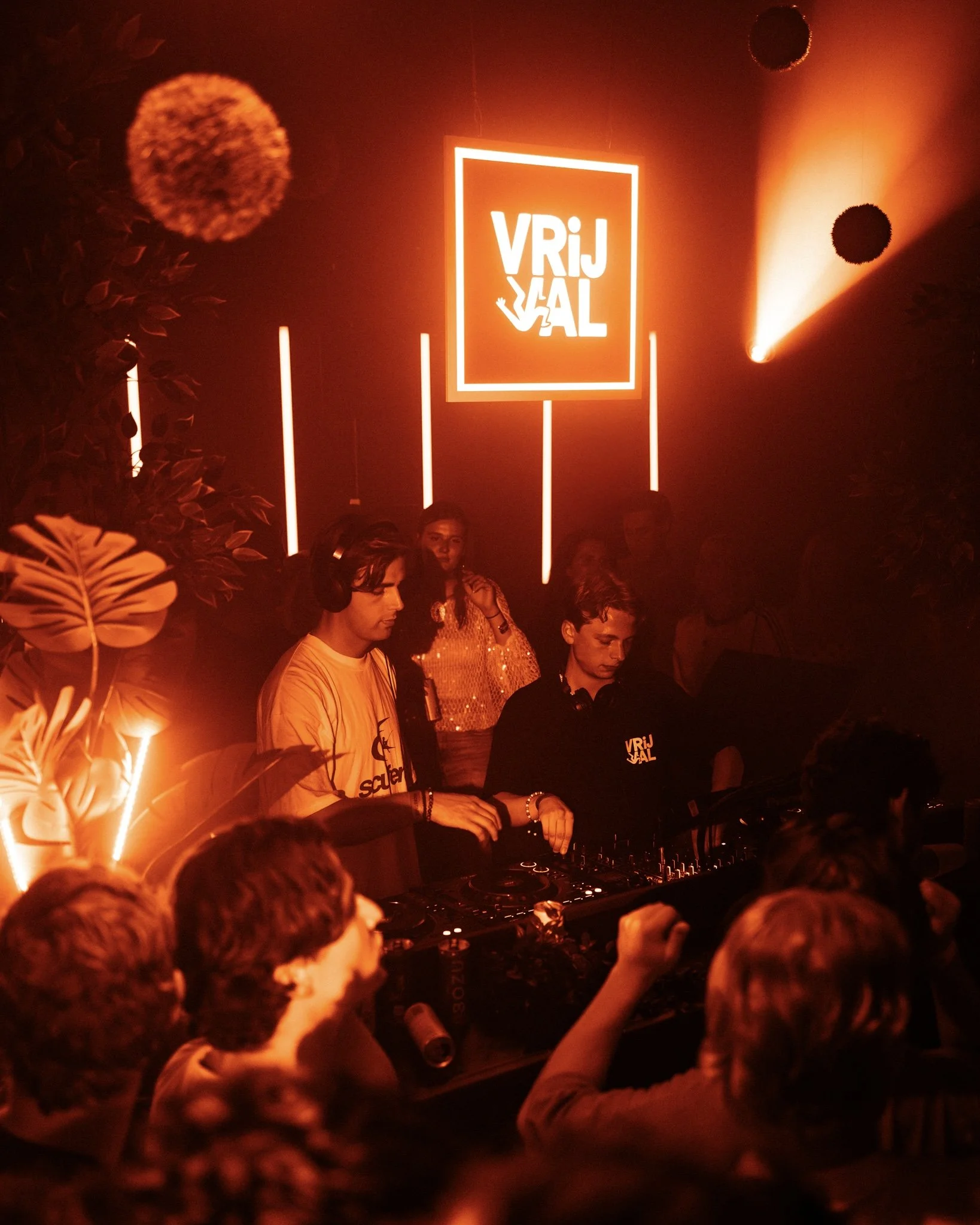 DJ performing at a night club with a glowing sign that says 'VRIJ is AL' illuminated in orange, with people dancing and enjoying the music in a vibrant, dark atmosphere.