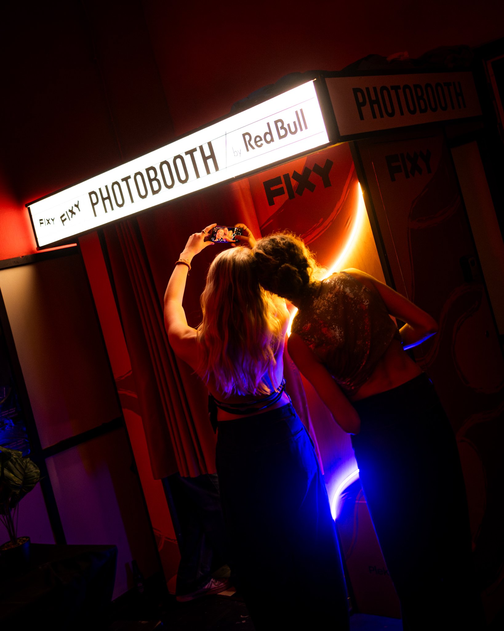 Two women taking a selfie together in front of a photo booth with bright signage and red and purple lighting.