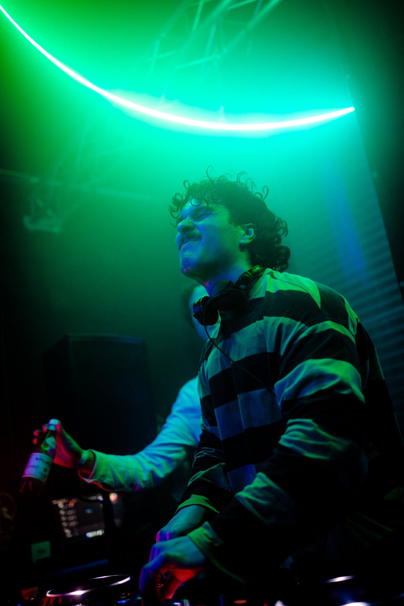 A DJ is performing at a nightclub, illuminated by green and blue neon lights, with headphones around his neck and a focused expression.