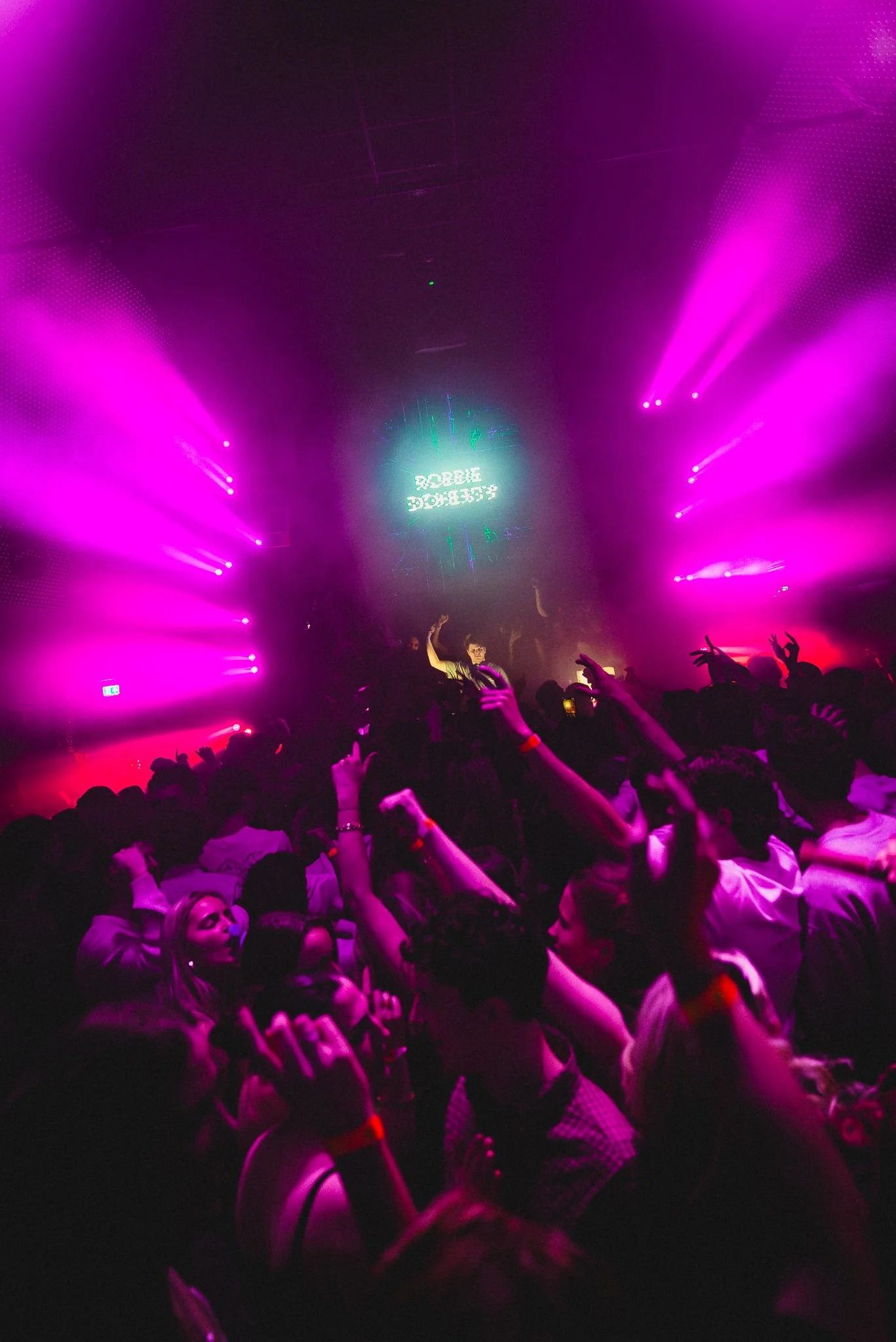 A crowded dance floor at a nightclub with purple and pink laser lights and a DJ performing on stage.
