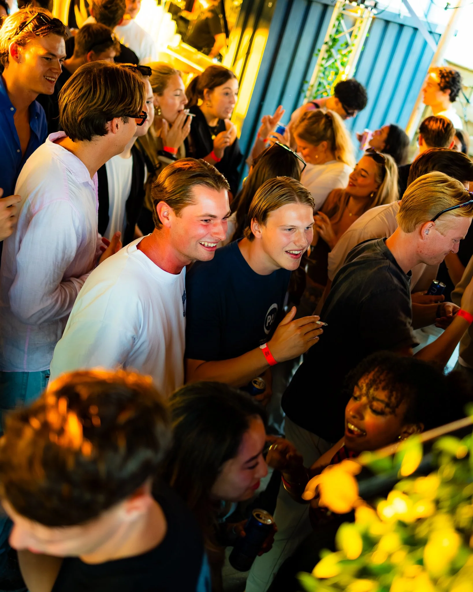 Crowd of young people at a lively social gathering or party, with some smiling and holding drinks, some engaged in conversation, and others taking photos, surrounded by colorful decorations.