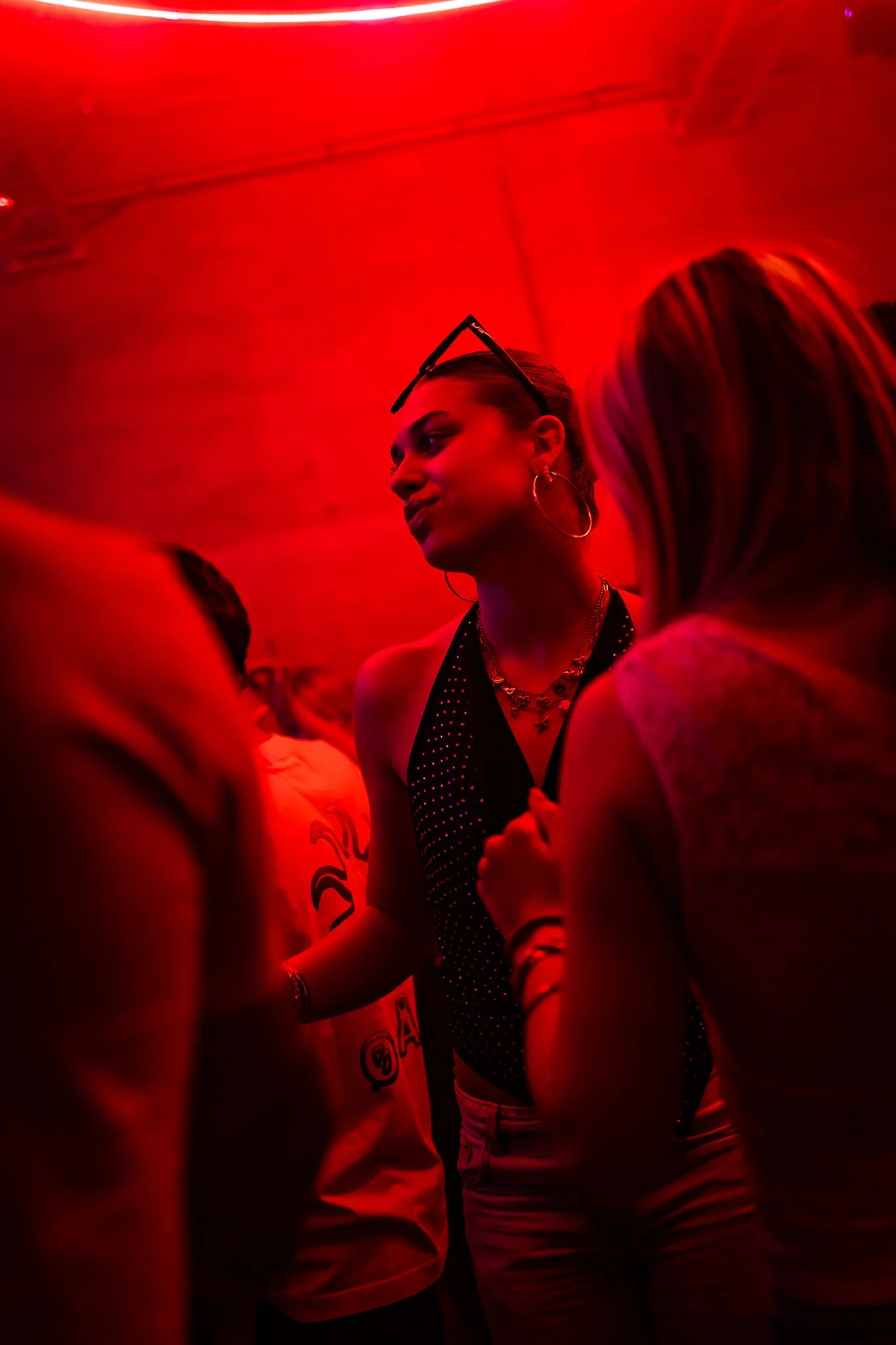 A woman with sunglasses on her head and hoop earrings is having a conversation in a dimly lit room with red lighting. She is wearing a black top with white polka dots and layered necklaces.