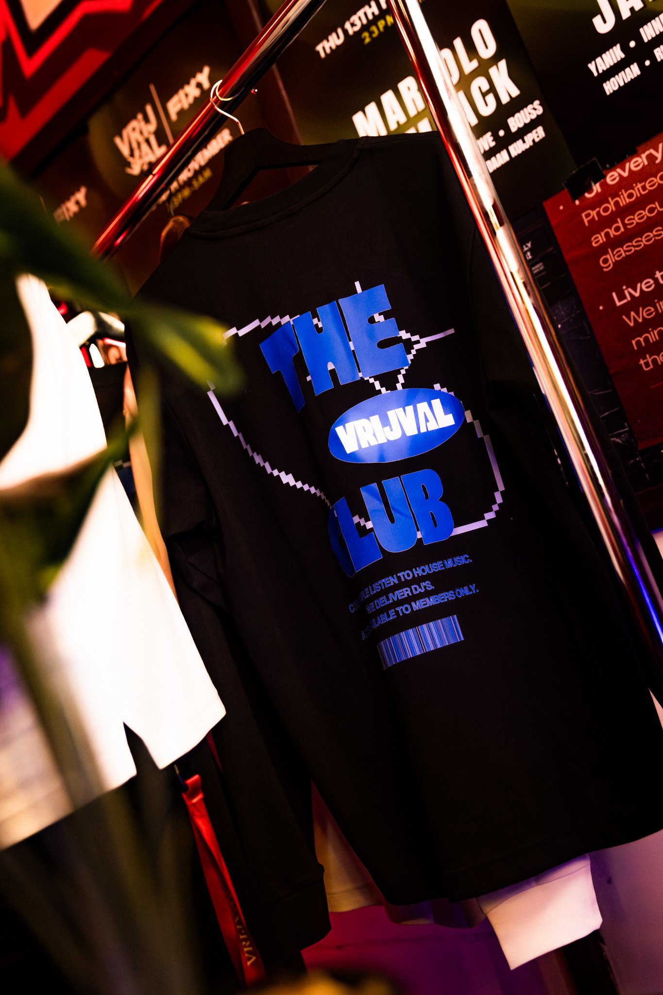 Black T-shirt with blue and white text reading 'The Virtual Club' hanging on a clothing rack in a dimly lit venue or club with red and black posters in the background.