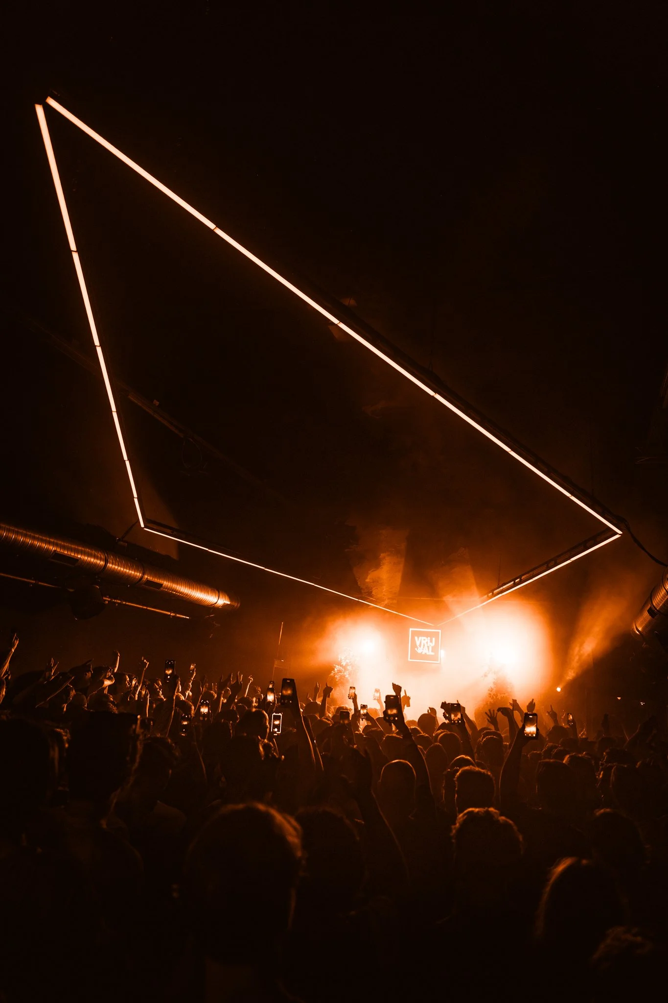 Crowd at a concert or music festival with many people holding up phones, illuminated by stage lights and orange lighting effects.