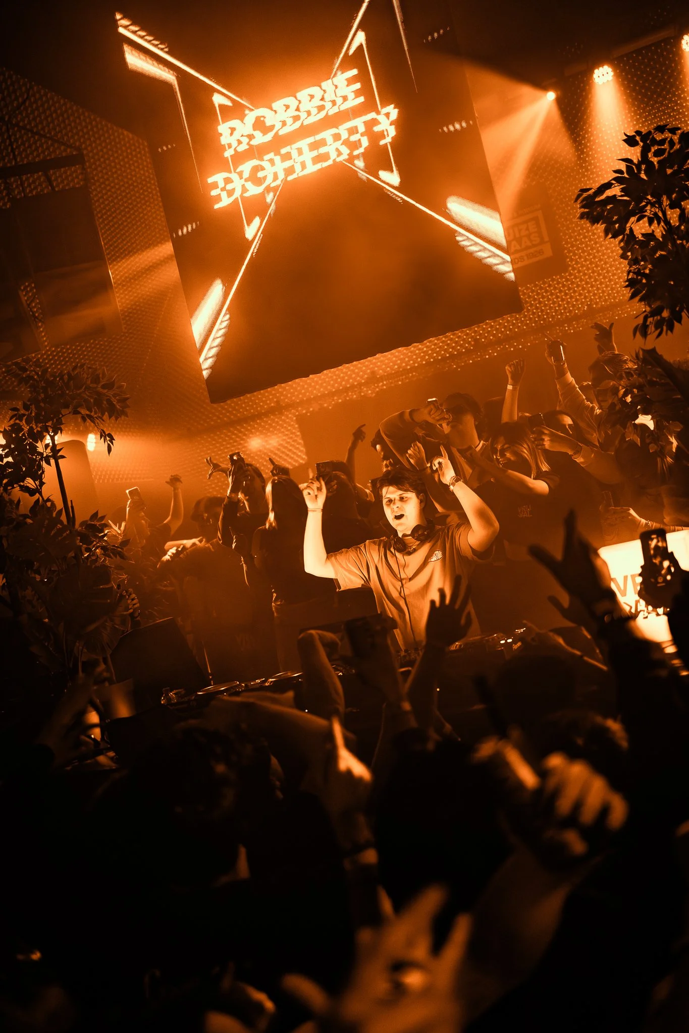 A DJ is performing at a nightclub with orange lighting. The crowd is dancing and raising their hands. A large screen displays the words 'Robbie Doherty' in neon-style font.