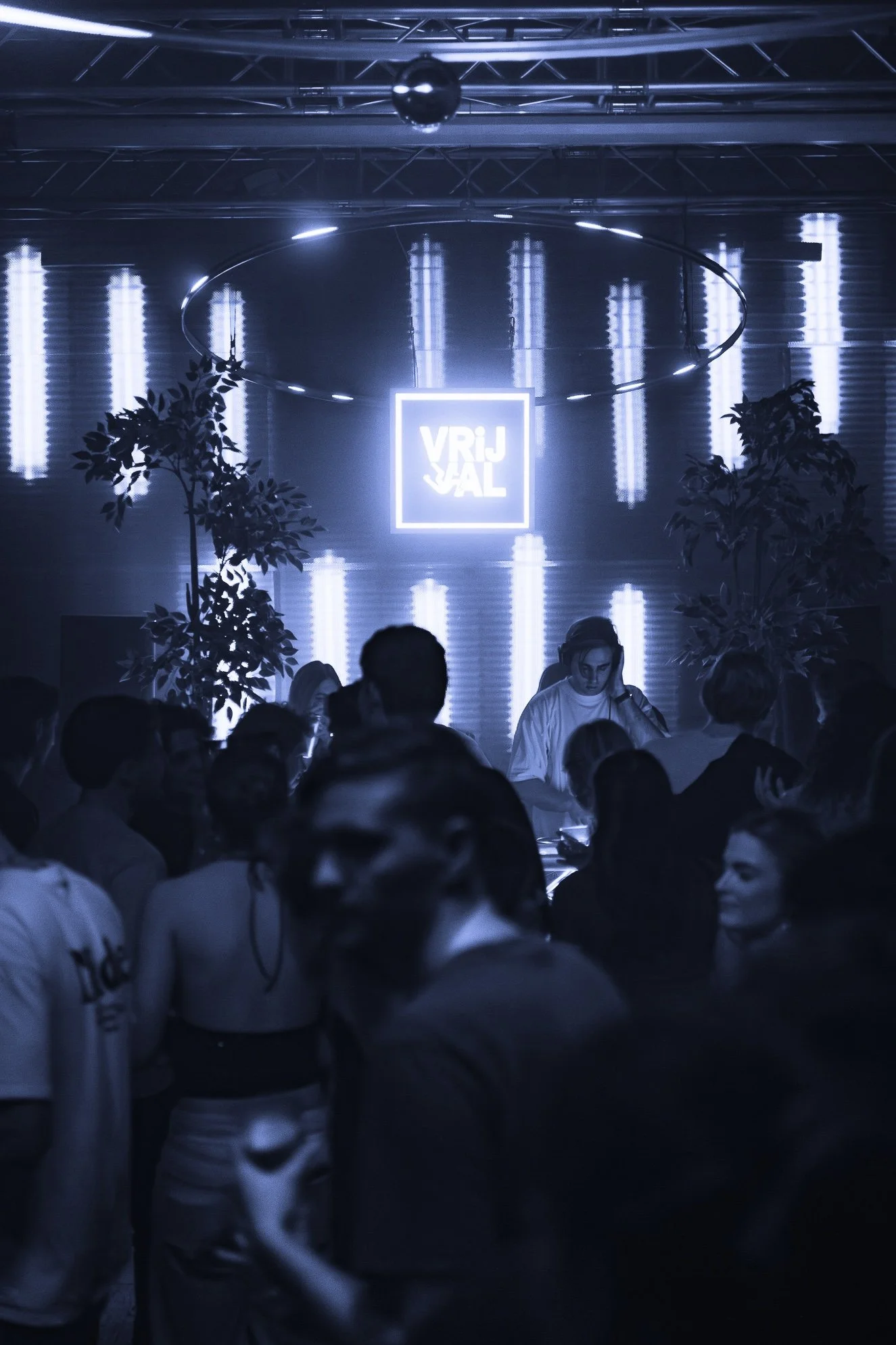 A nightclub scene with a DJ playing music on stage, illuminated by blue and white lighting, with a crowd of people dancing and socializing. There are two large plants on either side of the DJ, and a neon sign that reads "VRIJHAL" hanging above the stage.
