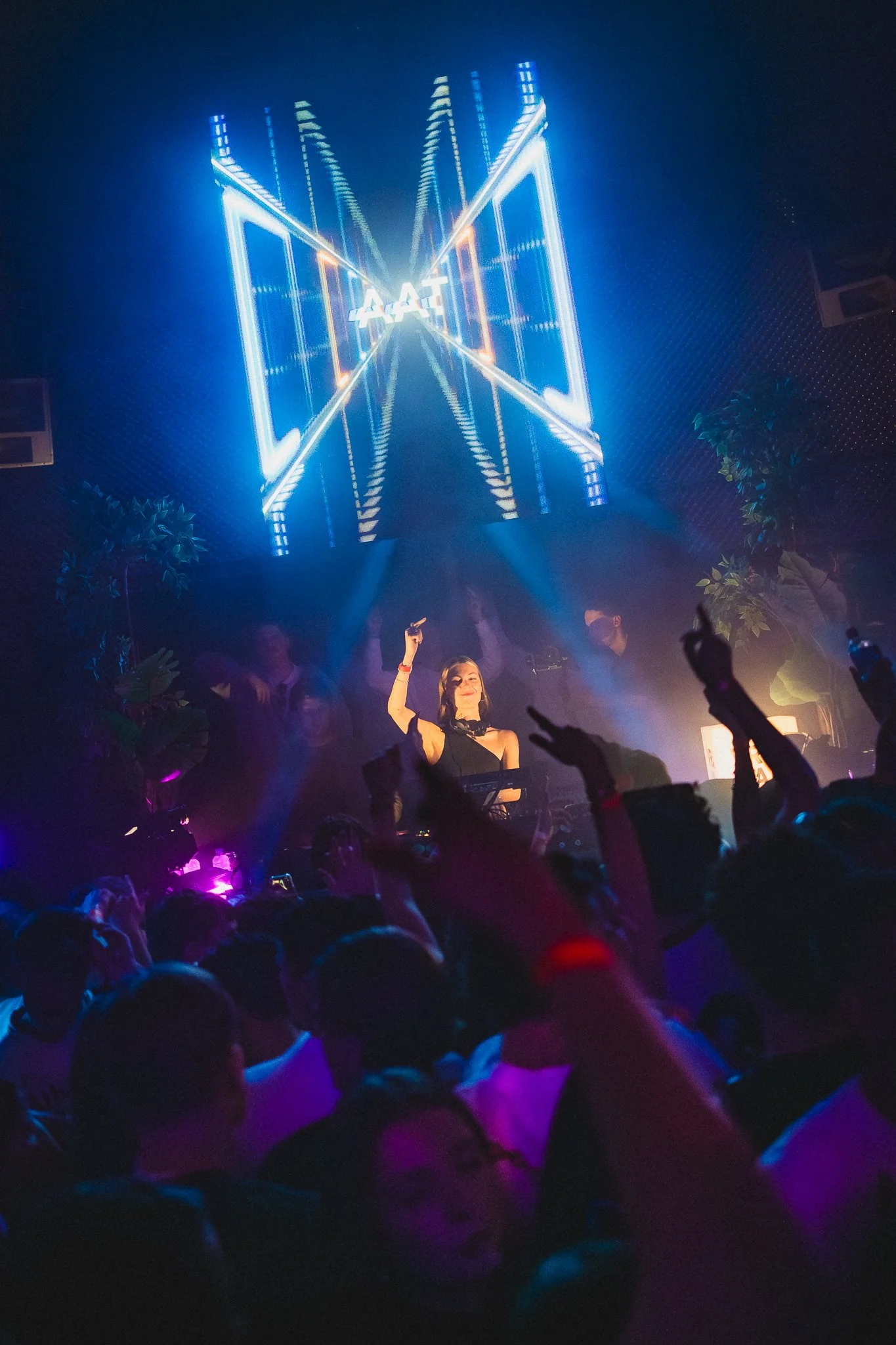 A woman DJ performs at a nightclub with blue LED light visuals on a large screen behind her, and an audience dancing in front.