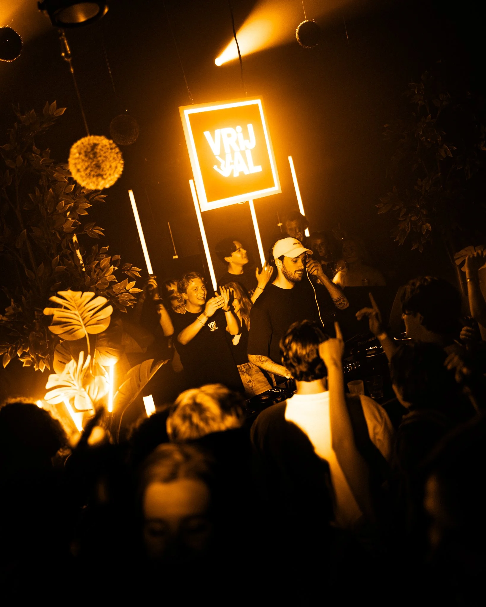 Crowd dancing and DJ playing at a nightclub with illuminated plants, neon sign reading 'VRIJJAL', and warm yellow lighting.