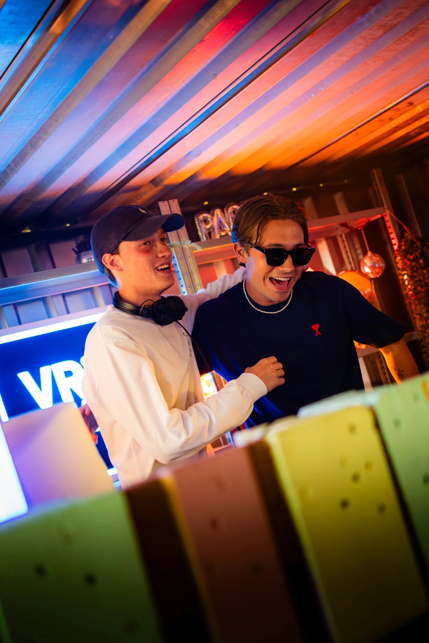 Two young men are celebrating together indoors with colorful lighting, wearing casual clothing and sunglasses, smiling and having fun.