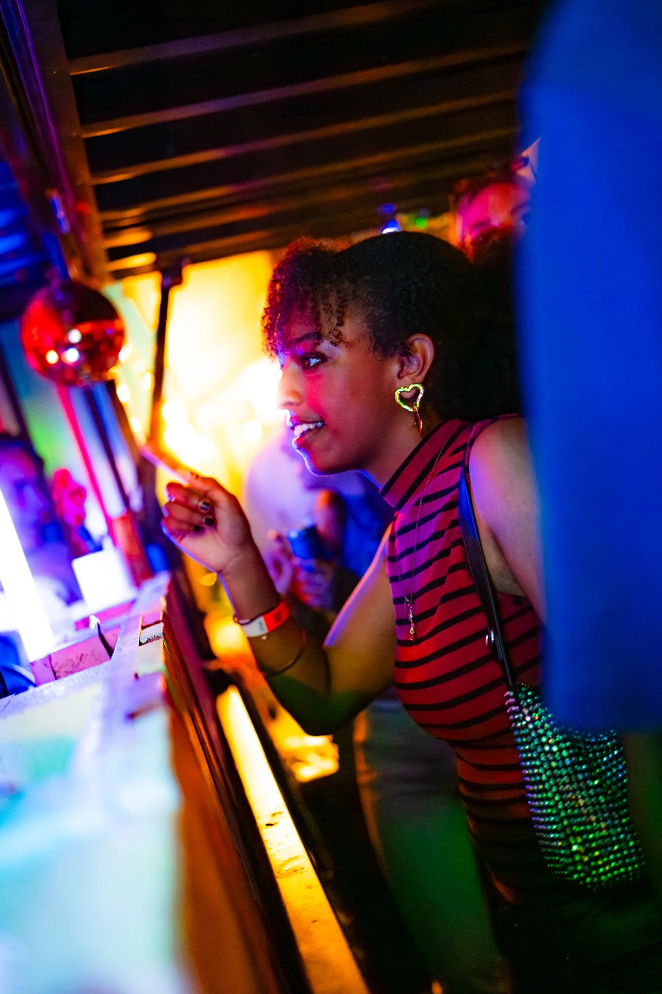 A woman with curly hair and heart-shaped earrings smiling and talking to someone at a bar in a colorful, dimly lit nightclub.