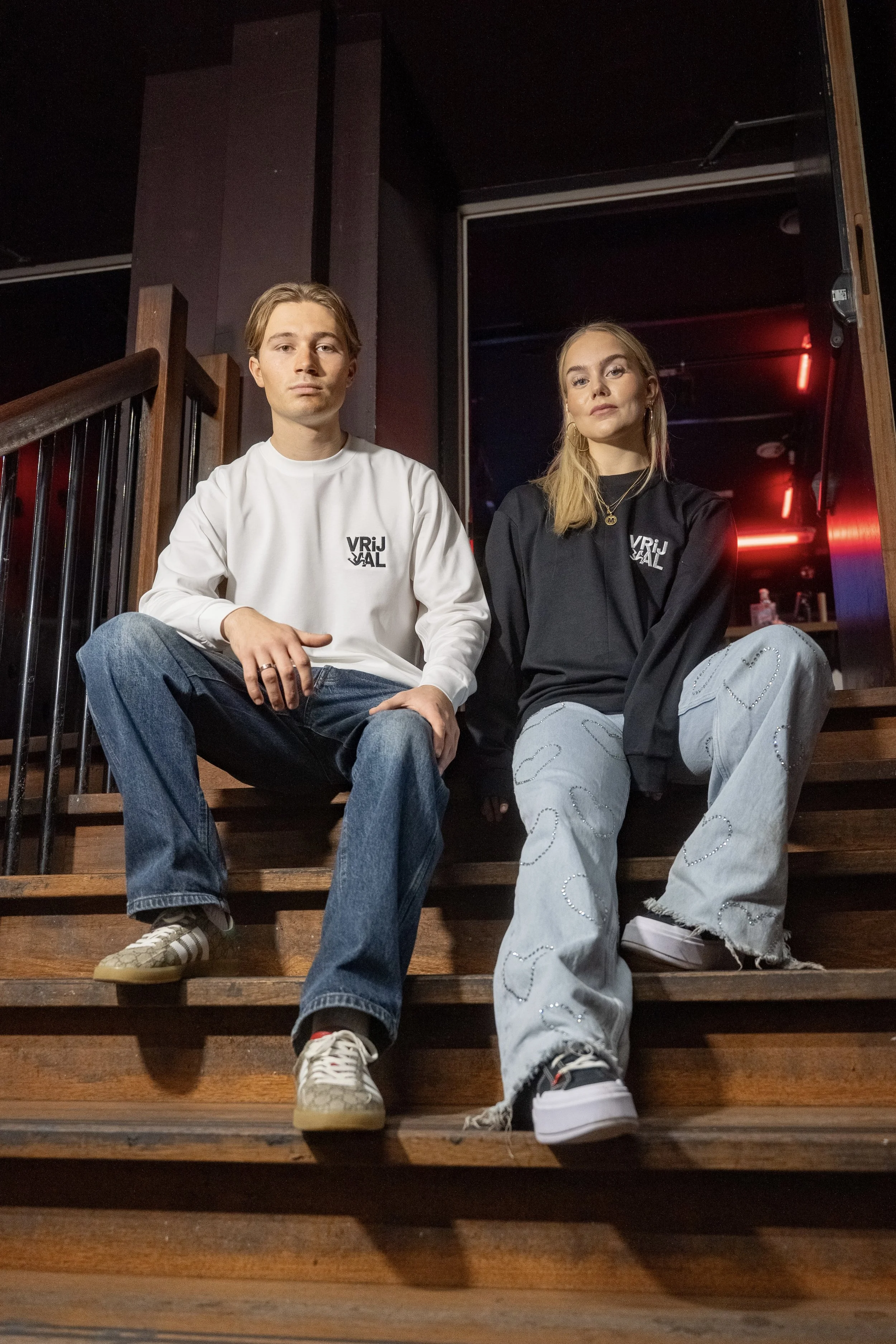 A young man and woman sitting on wooden stairs in a dimly lit indoor space. Both are casually dressed, with the man in a white long-sleeve shirt and jeans, and the woman in a black sweatshirt and ripped jeans. They have serious expressions.