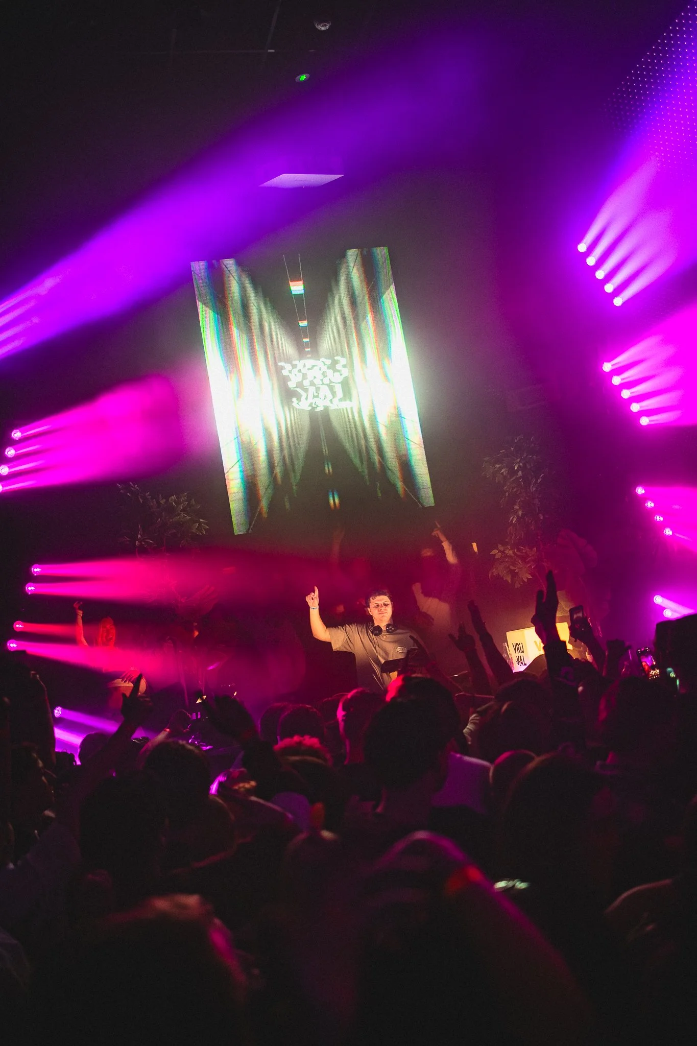 A DJ performing at a concert with purple and pink stage lighting. The audience is raising their hands and taking photos.