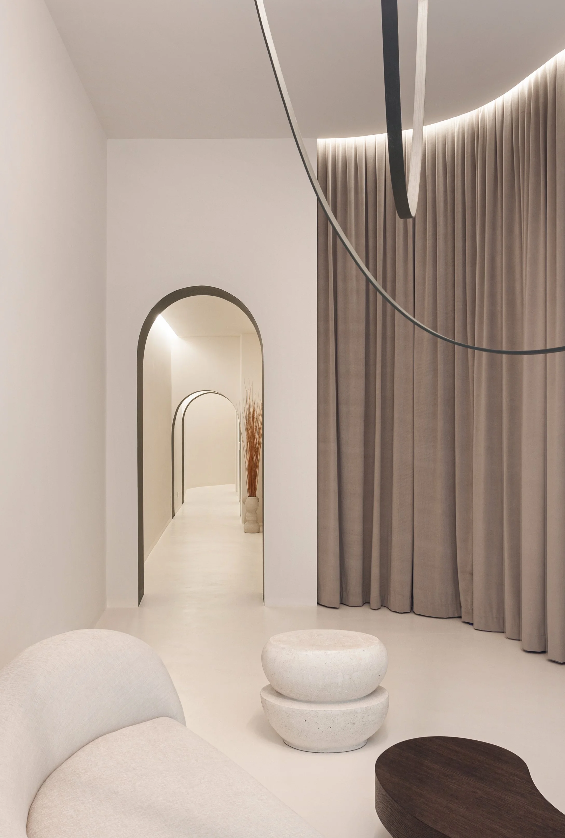Interno minimalista con divano beige, appendiabiti di pietra, tende beige, e di archi nel corridoio.