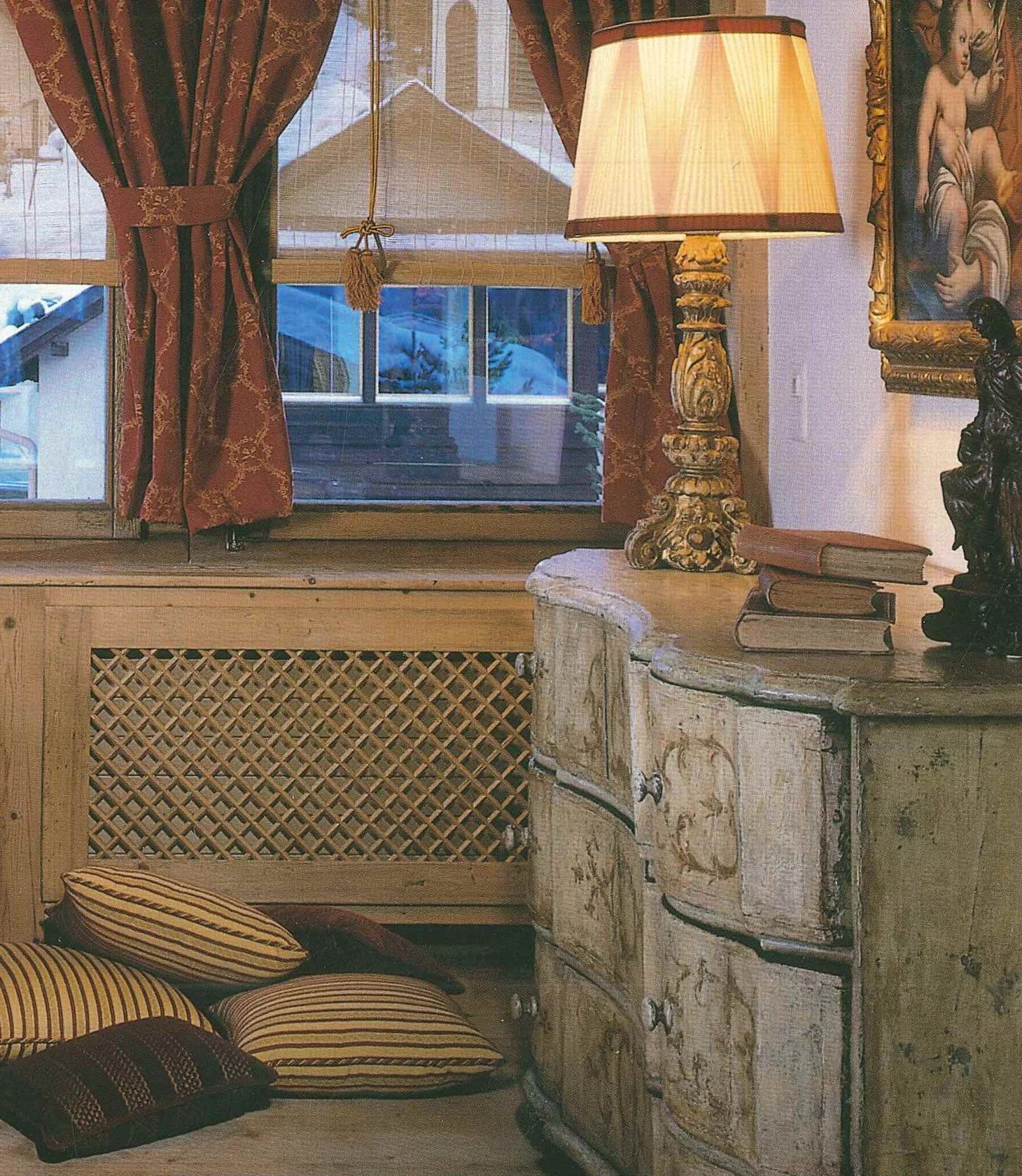 Interno di una stanza con una finestra, tende rosse e parapetto in legno; sul tavolo c'è una lampada e alcuni libri. Di fianco un mobile antico con mobiletto decorato, e sul pavimento ci sono cuscini con fantasie a righe.