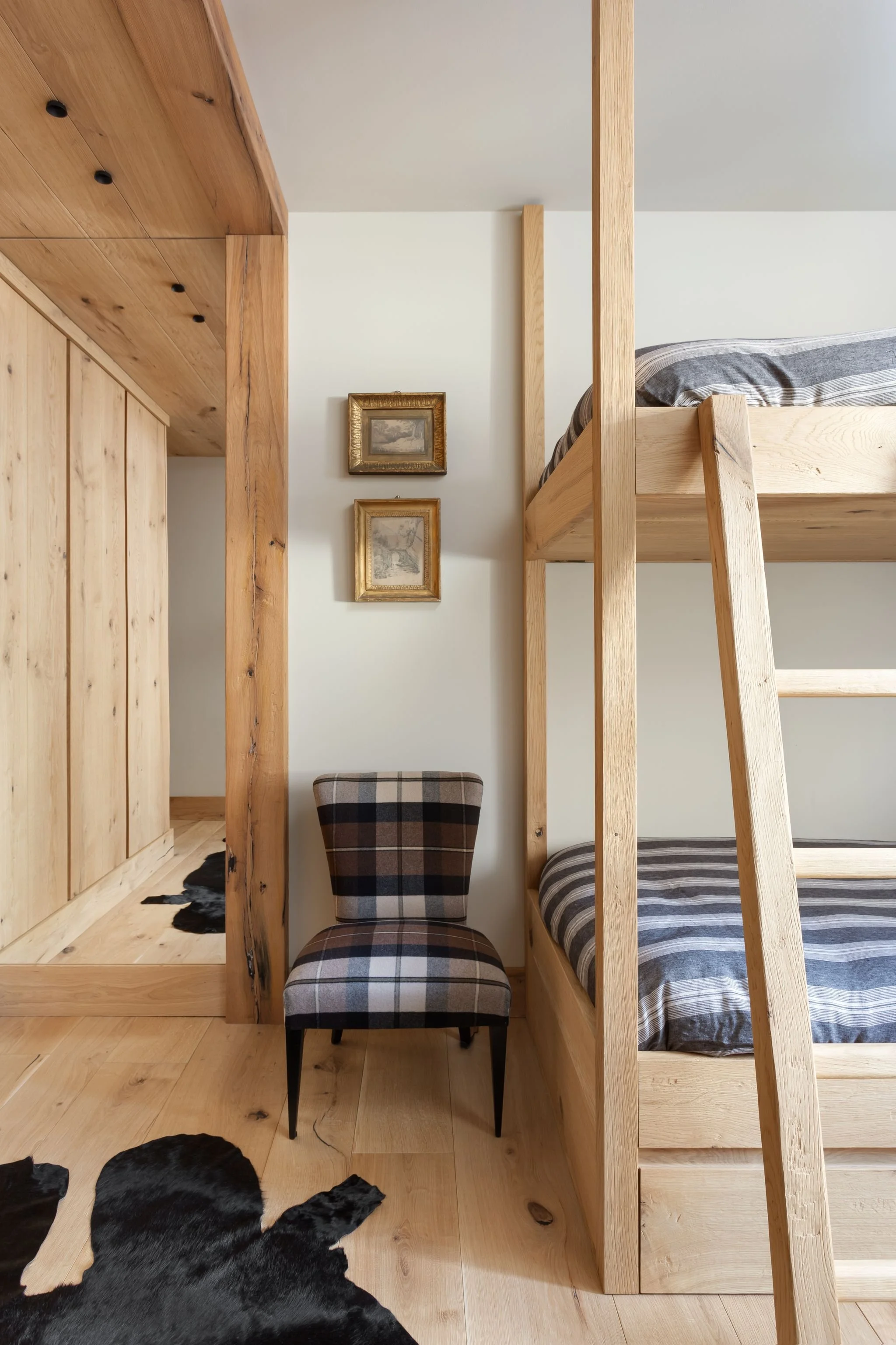 Sala con letto a castello in legno, sedia con tessuto a scacchi, quadri dorati, tappeto di pelle nera, pareti bianche e pavimento in legno.