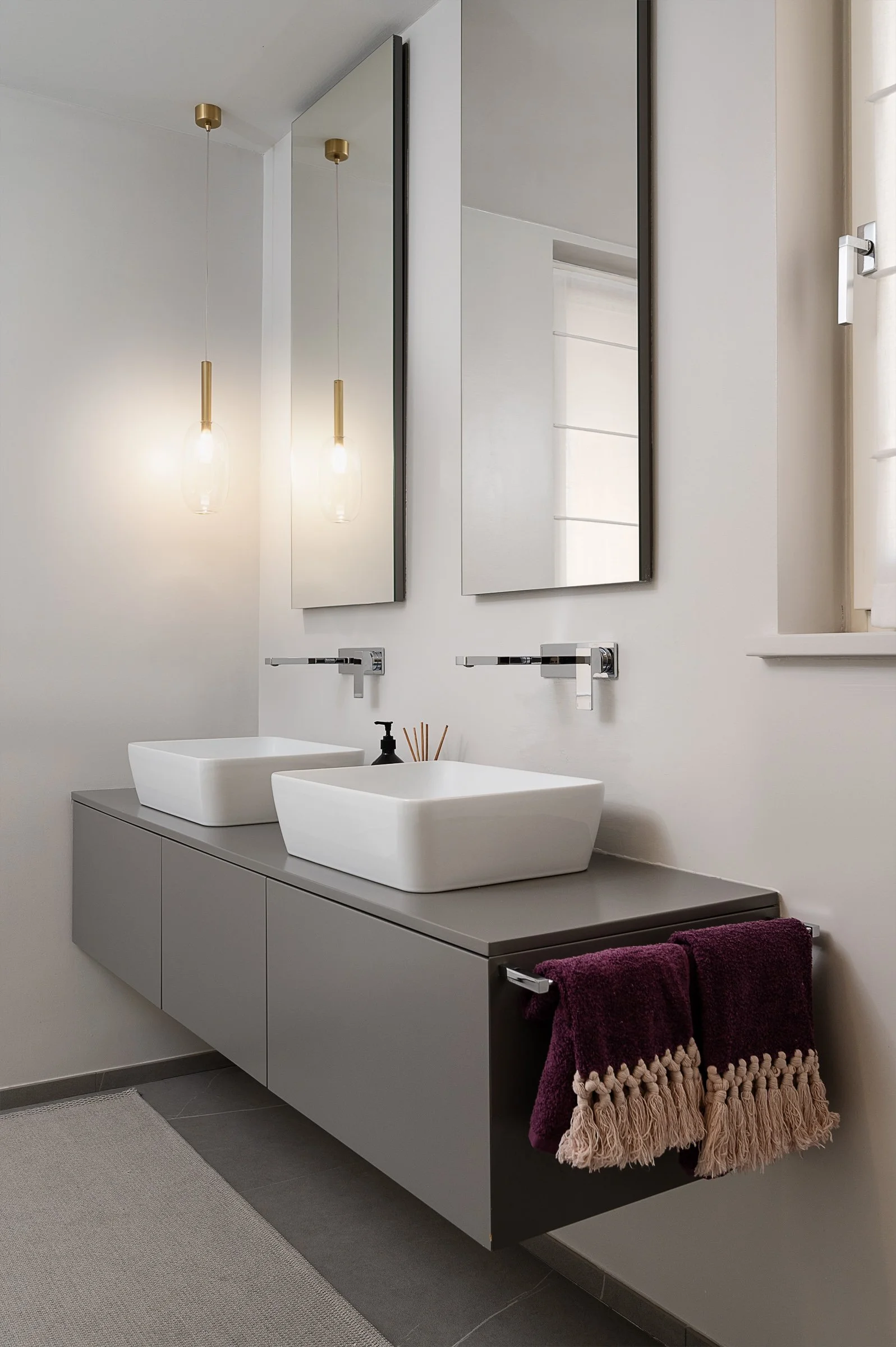 Bagno moderno con due lavabi bianchi a scaletta su un mobile grigio, specchi a tutta altezza e due lampade pendenti dorate.