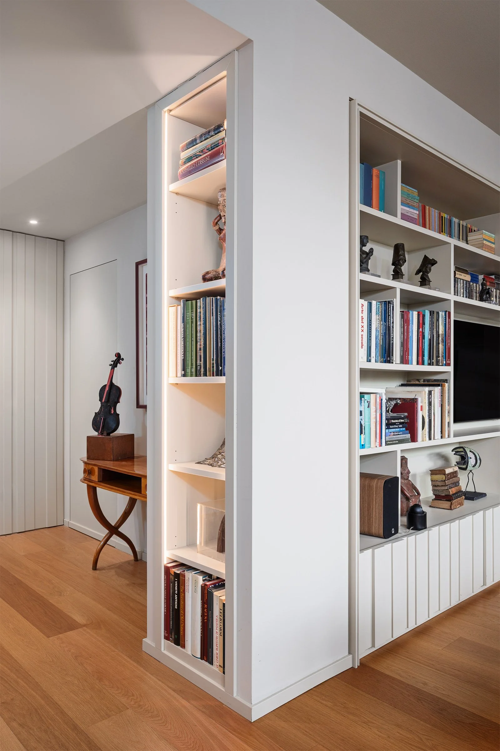 Angolo di una stanza con librerie bianche piene di libri e statue decorative, pavimento in legno e pareti bianche.
