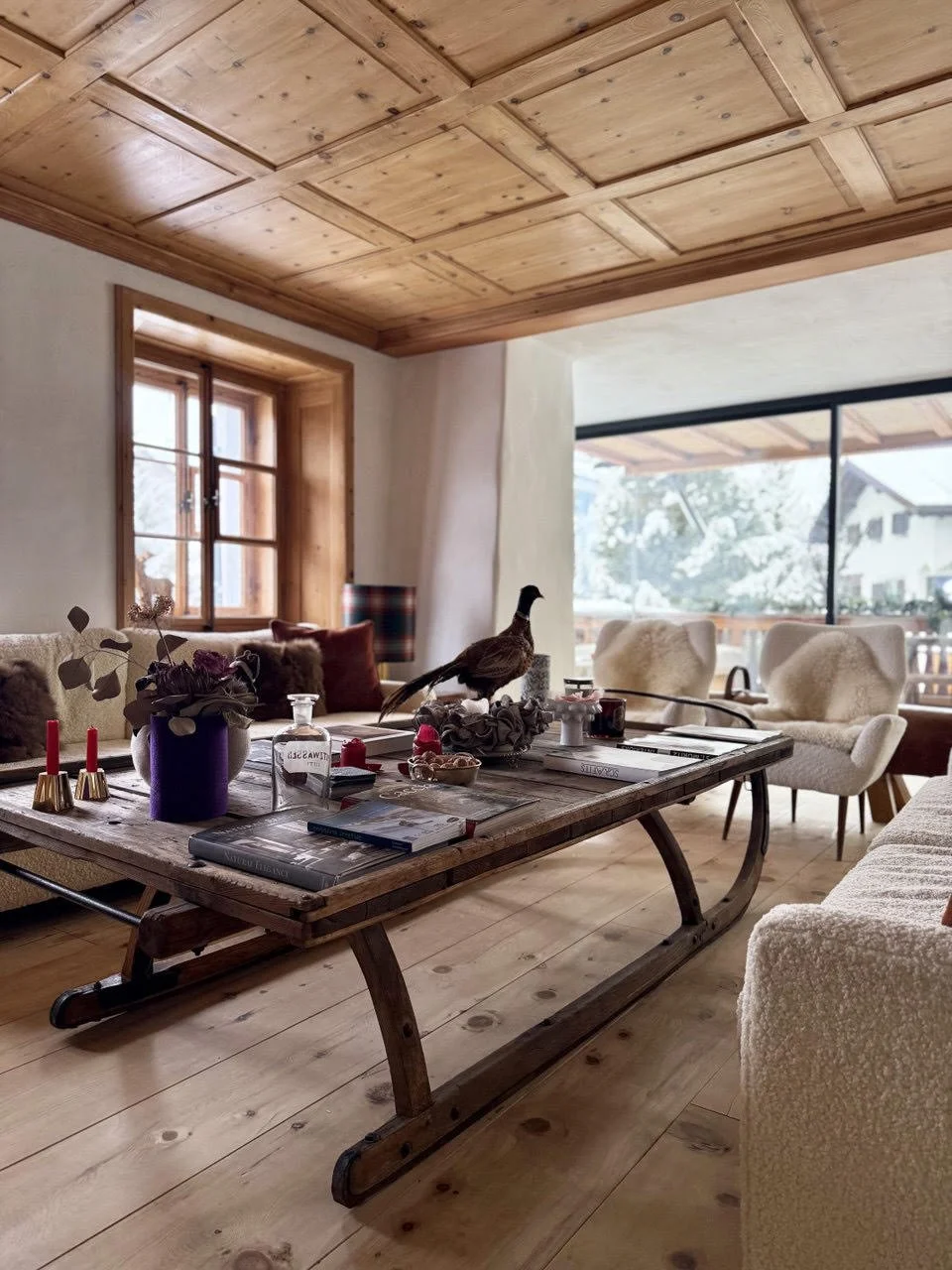 Interno di una stanza con soffitto in legno, grande finestra e mobilio in stile rustico. C'è un tavolo con vari oggetti decorativi e una statua di un uccello. Sedie e poltrone in tessuto morbido, con vista su uno slargo esterno con case e alberi.