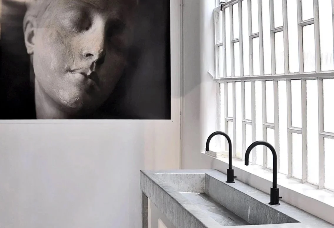 Interno di un capannone con pareti di vetro, lavandino in pietra e un grande quadro con un volto di donna in bianco e nero