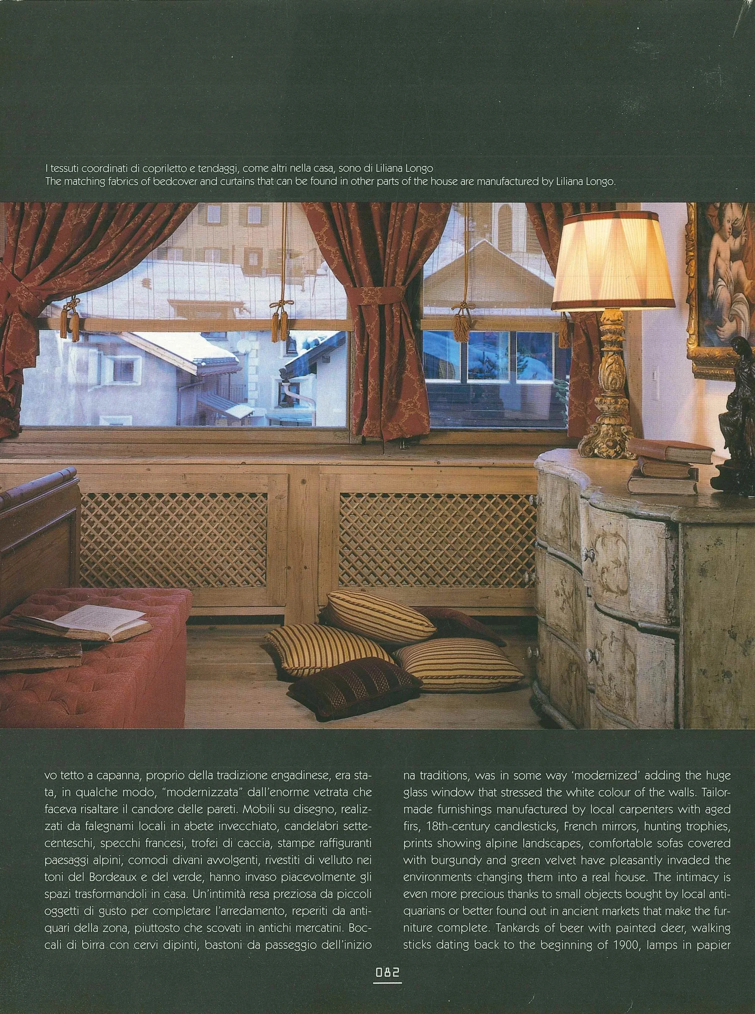 Interno di una stanza con tende rosse, cuscini e mobili in stile antico, con una finestra che mostra case con tetti innevati.