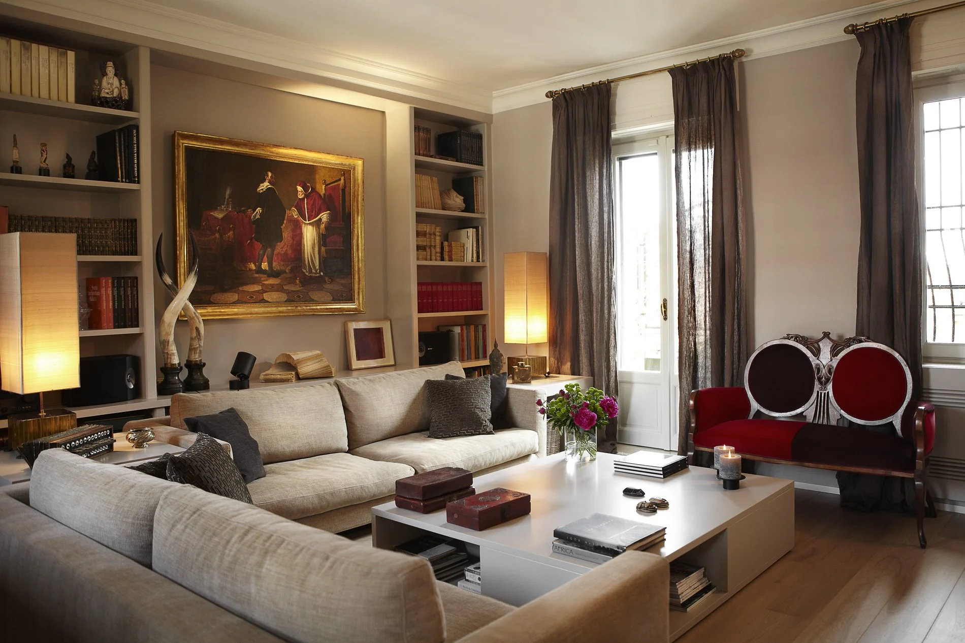 Salotto elegante con divani beige, libro e decorazioni su tavolino e parete con quadri, librerie piene di libri, lampade e tende beige e marroni.