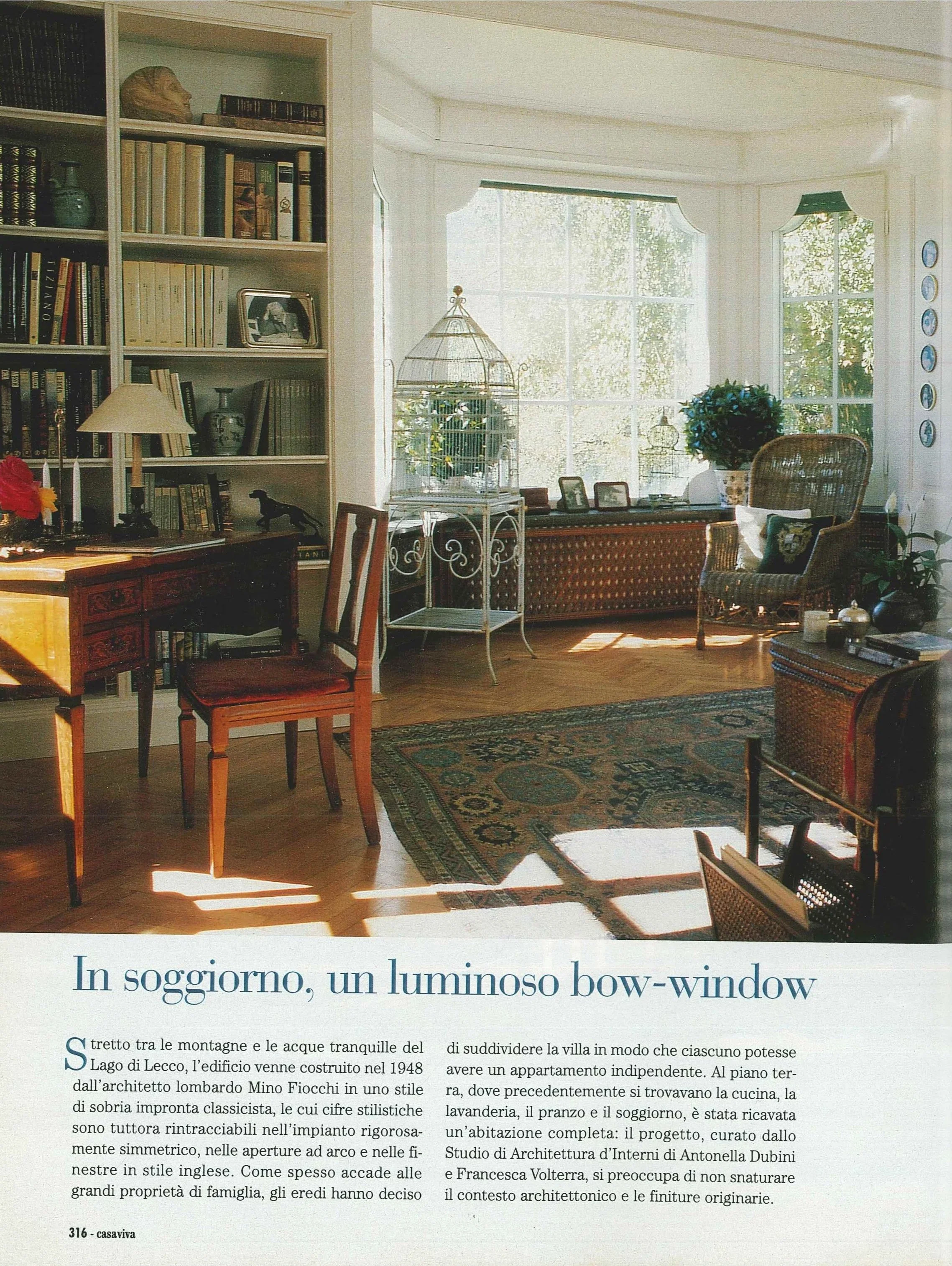 Interior di soggiorno con grande finestra luminosa, mobili in legno, scaffali pieni di libri, un tavolo con una sedia, divano, poltrona e piante.