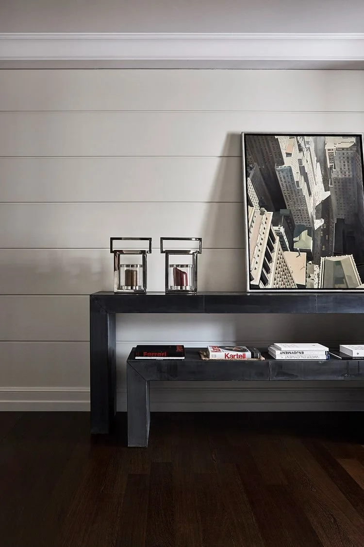 Una console nera con libri e due candelieri di vetro sulla superficie, un quadro appoggiato contro la parete con immagine di un grattacielo