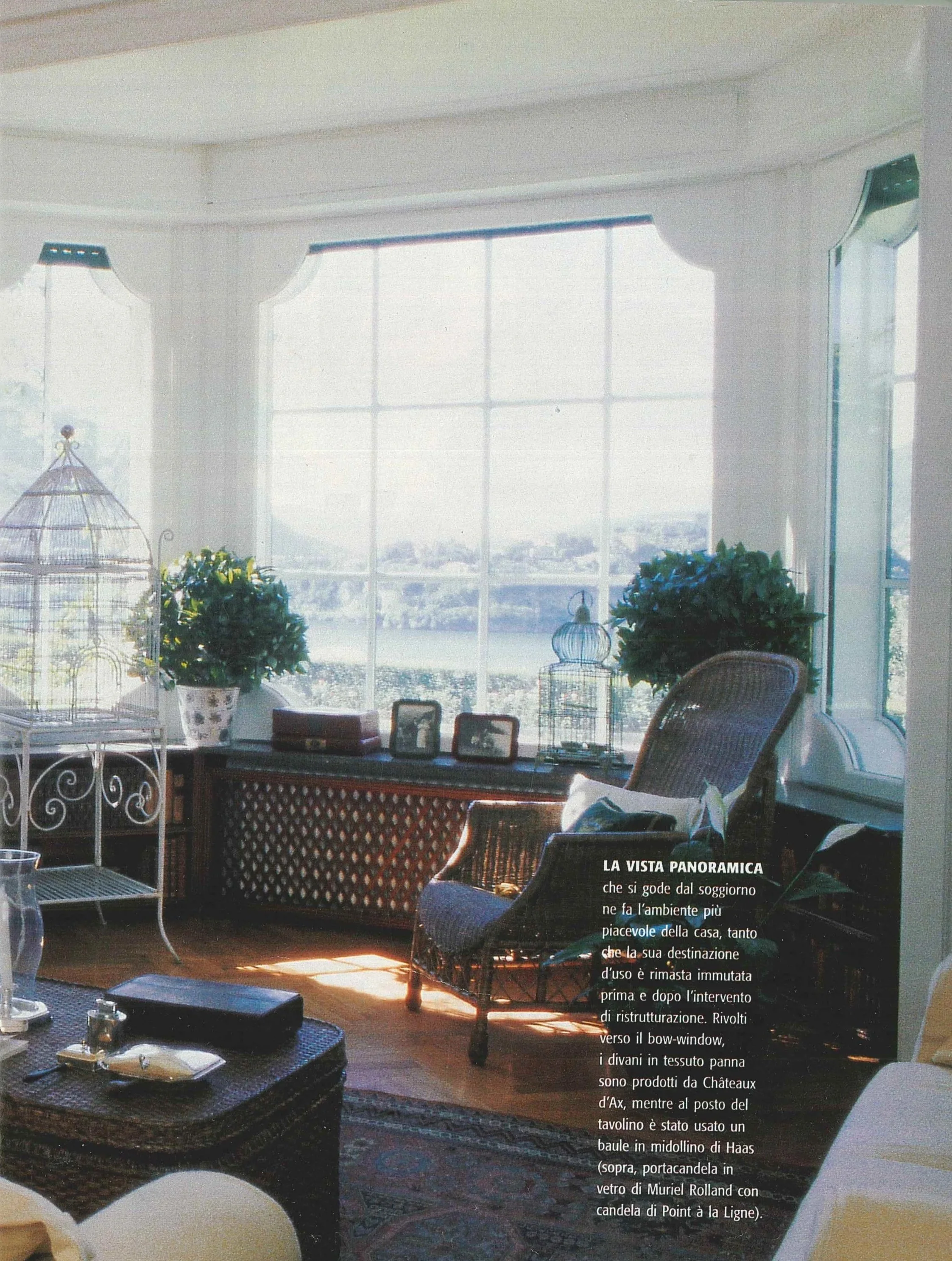 Interno di una stanza con grandi finestre panoramiche, piante verdi, sedie, cassetti, e oggetti decorativi, con vista su un paesaggio esterno.