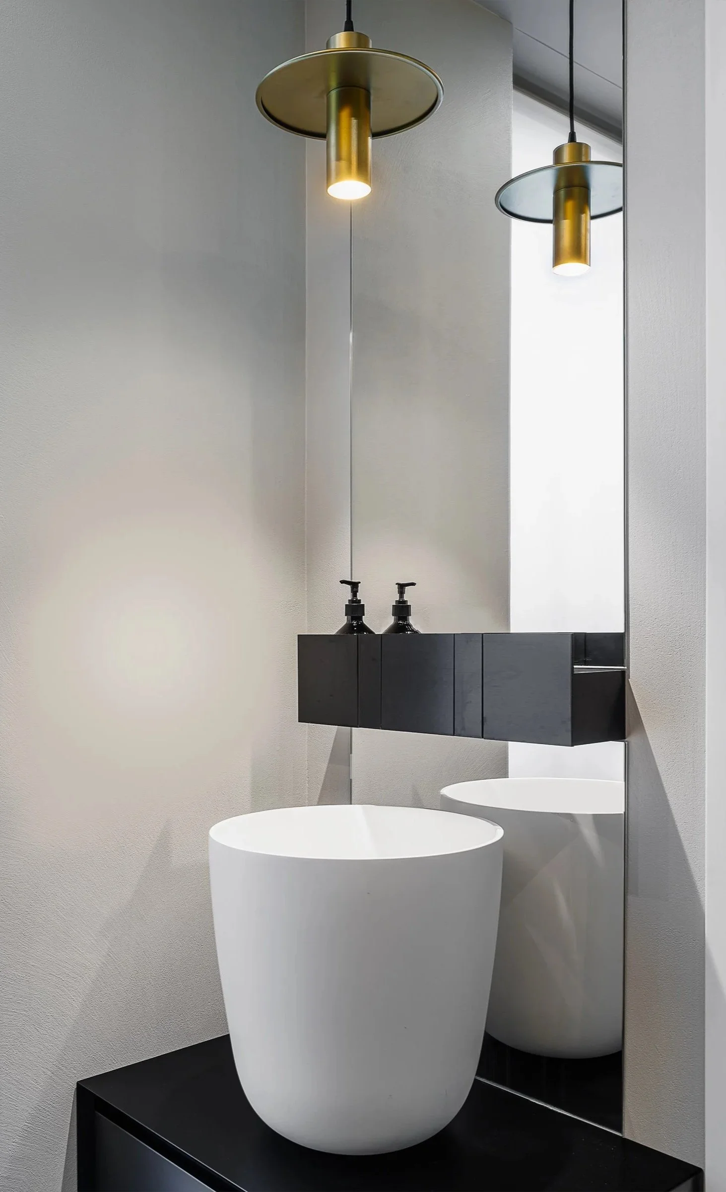 Lavabo bianco rotondo con un grande specchio e due distributori di sapone neri sopra un mobile nero. Due lampade pendenti dorate appese dal soffitto.