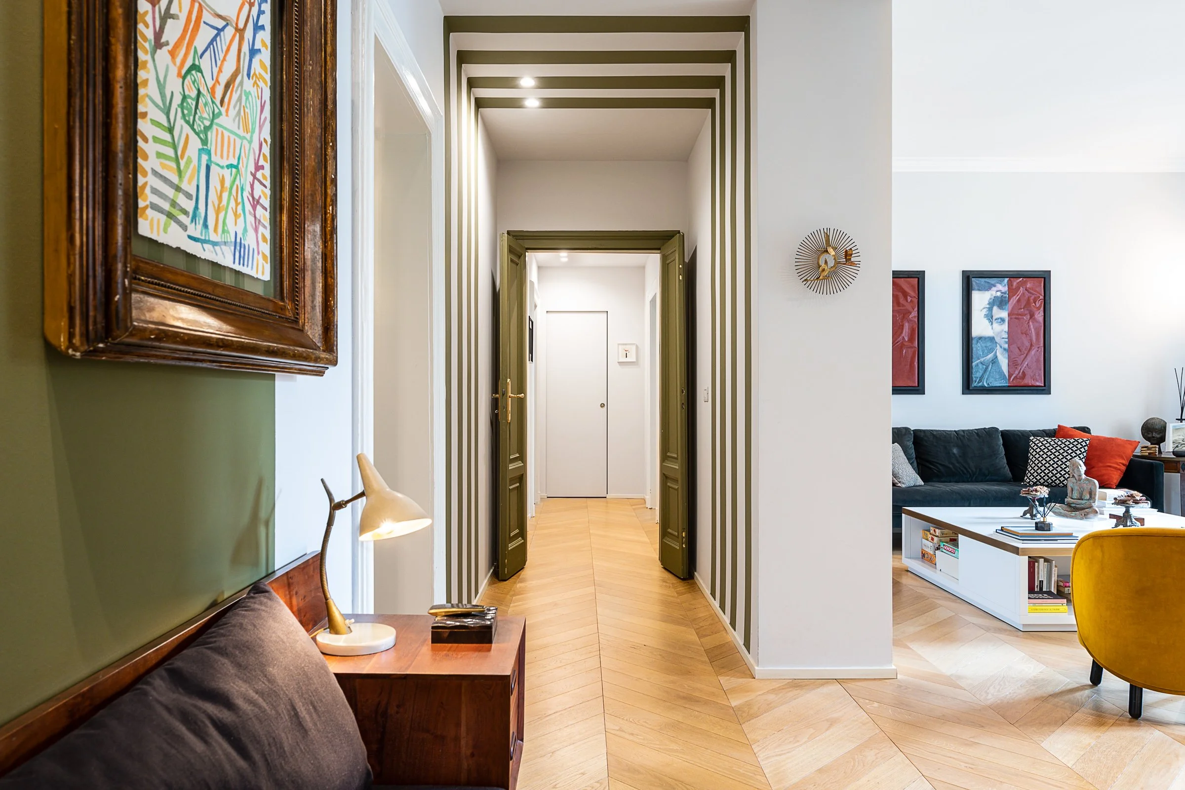 Interno di un appartamento con un corridoio centrale, pareti con righe verdi e bianche, pavimento di legno, e in fondo una porta bianca. Sul lato destro, un soggiorno con divani e quadri alle pareti, e a sinistra una zona con una lampada da tavolo e quadro.