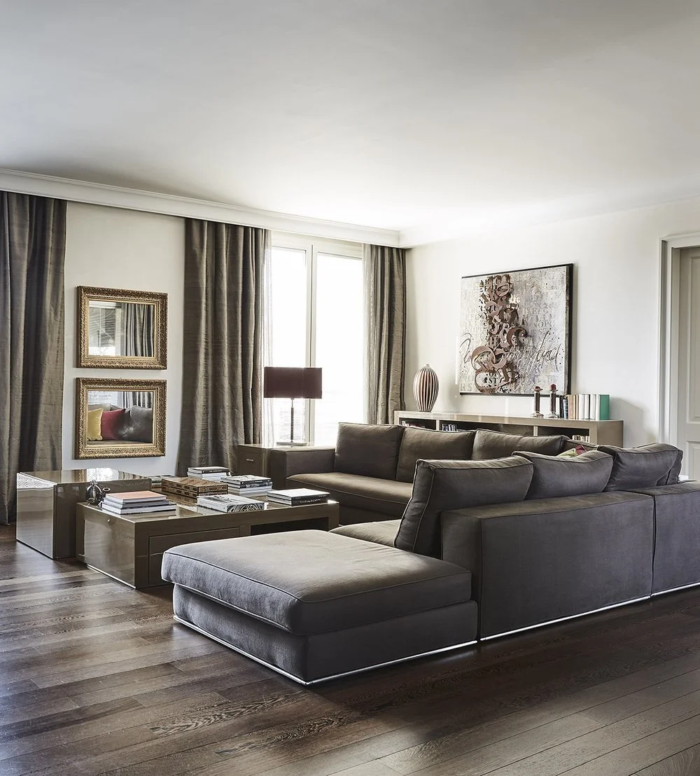 Soggiorno con divano grigio, tappeto, tavolini con libri, pareti con quadri e specchi, lampada da tavolo, e decorazioni.