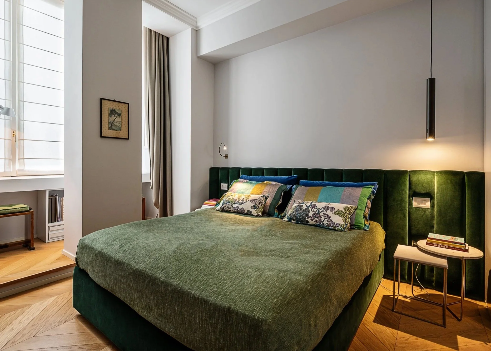 Camera da letto moderna con letto verde, cuscini colorati, applicazioni a parete e una piccola consolle con libri.