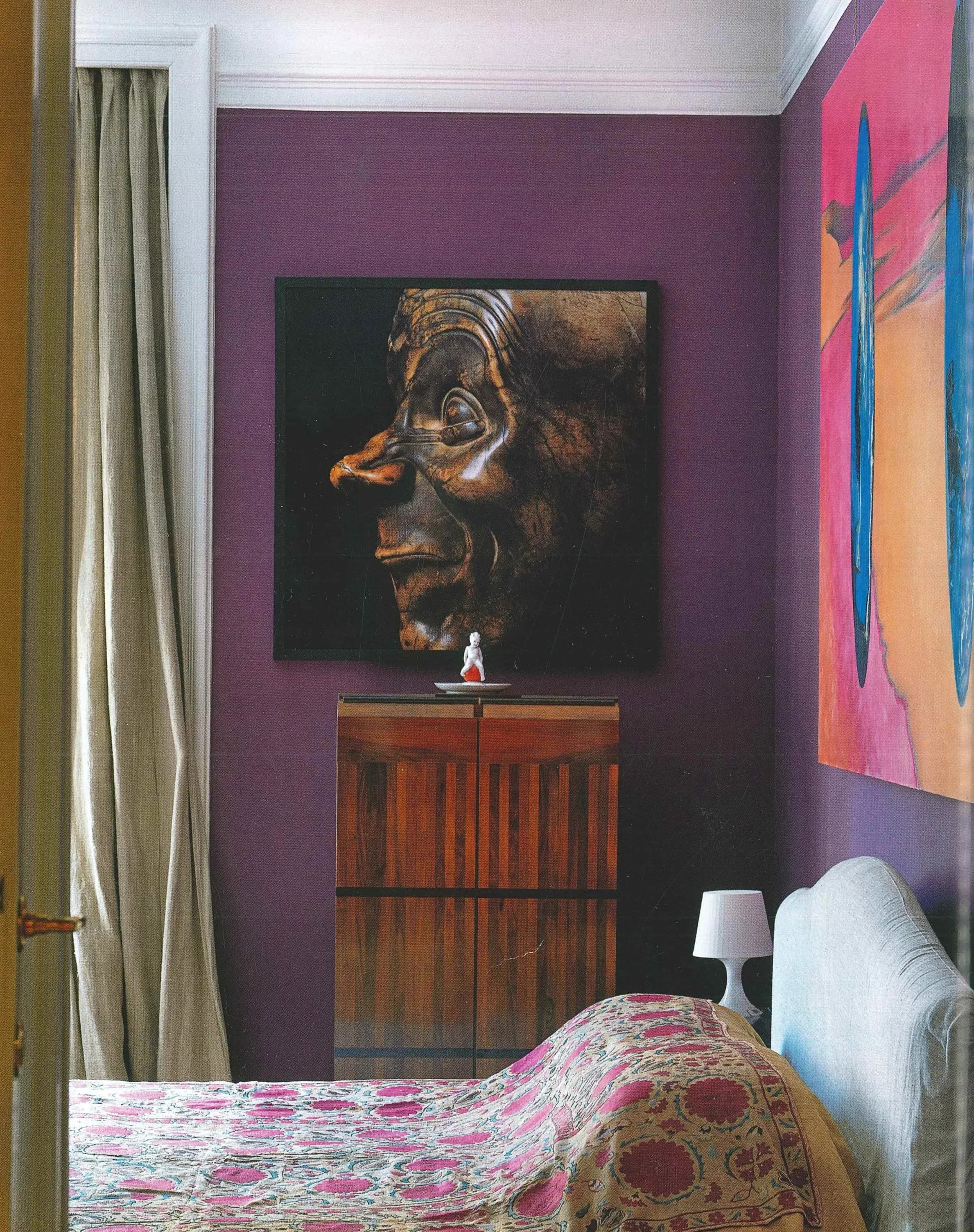 Interno di una stanza con pareti viola, un letto con copertura colorata, un comodino con lampada, un quadro di un volto stilizzato sull'armadio e un'altra grande opera d'arte sulla parete.