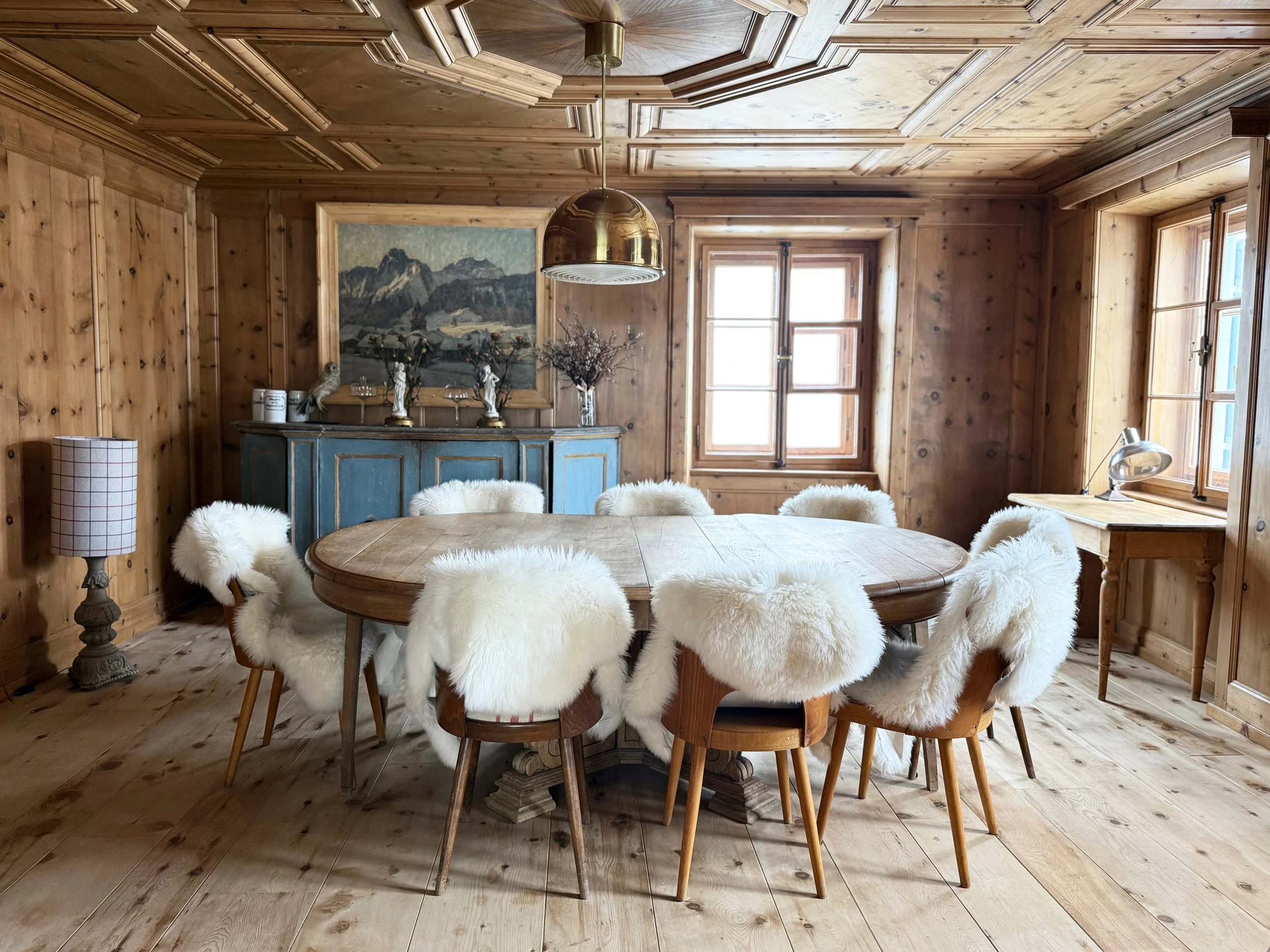Una stanza in stile rustico con pareti e pavimenti in legno, tavolo rotondo in legno circondato da sedie con poggiabraccia in pelliccia bianca. Sullo sfondo, una credenza blu con decorazioni e un quadro con montagne. Due finestre lucide illuminano la stanza, una scrivania con lampada in stile industriale e un lampione con lampada a stelo. decorazioni e vasi con ramoscelli.