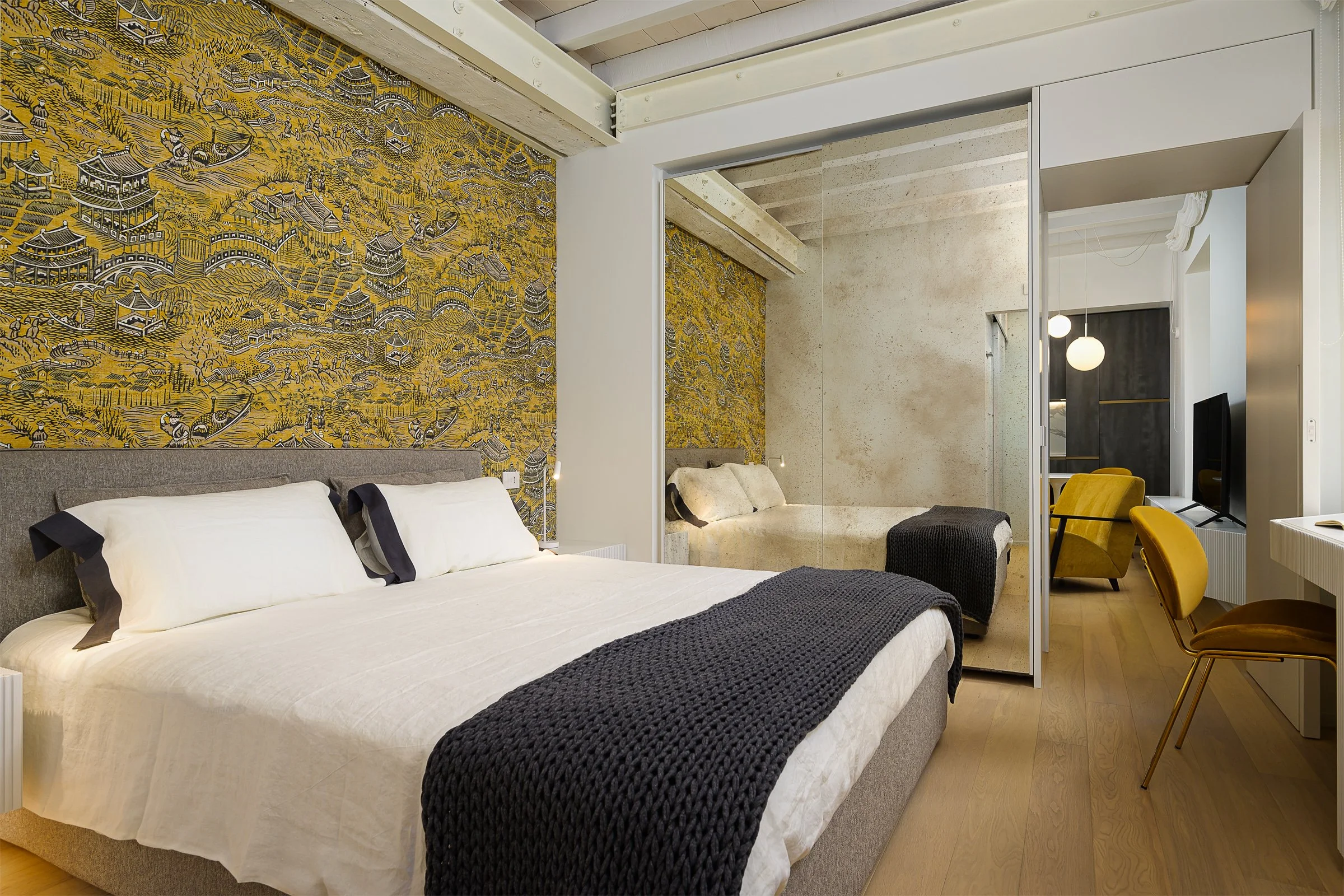 Camera da letto moderna con parete decorata con murale giallo e nero, letto con biancheria bianca e coperta nera, area riservata con divano beige e sedie gialle nello sfondo.