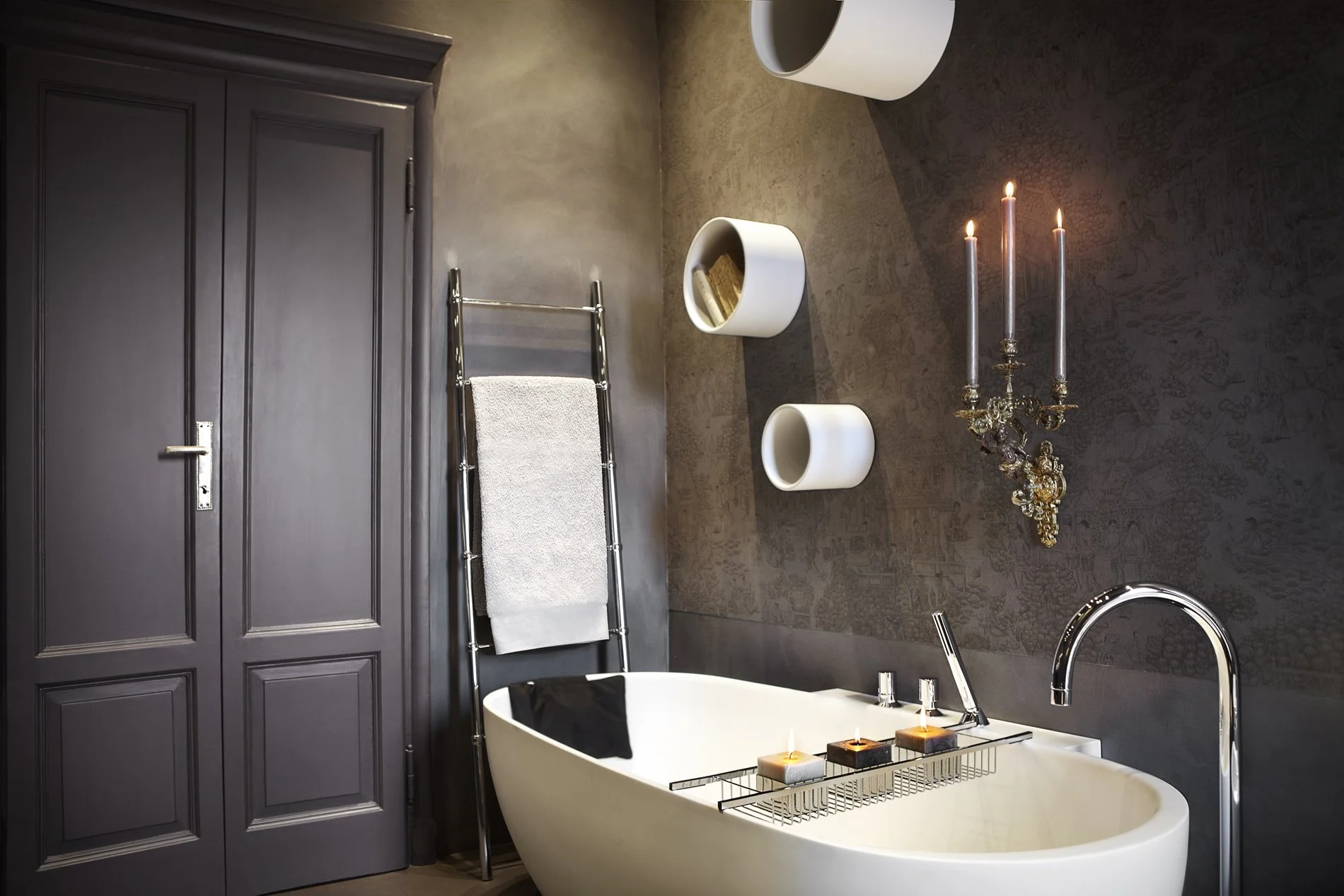 Bagno elegante con vasca da bagno bianca, candela accesa sulla vasca, lampadario d'epoca con candele accese, pareti scure con decorazioni, porta grigia e scaffale con asciugamani.