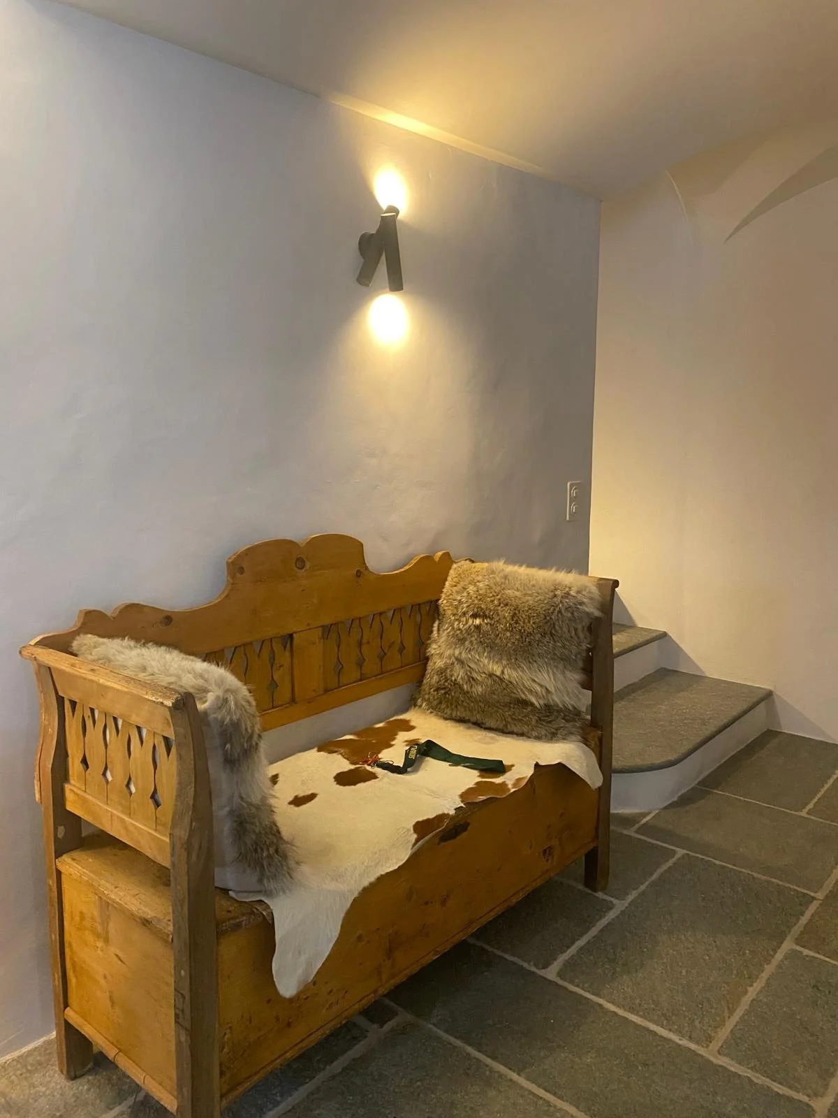 Seduta di legno decorata con pelliccia e cuscini, in un angolo con pareti chiare e lampada a soffitto